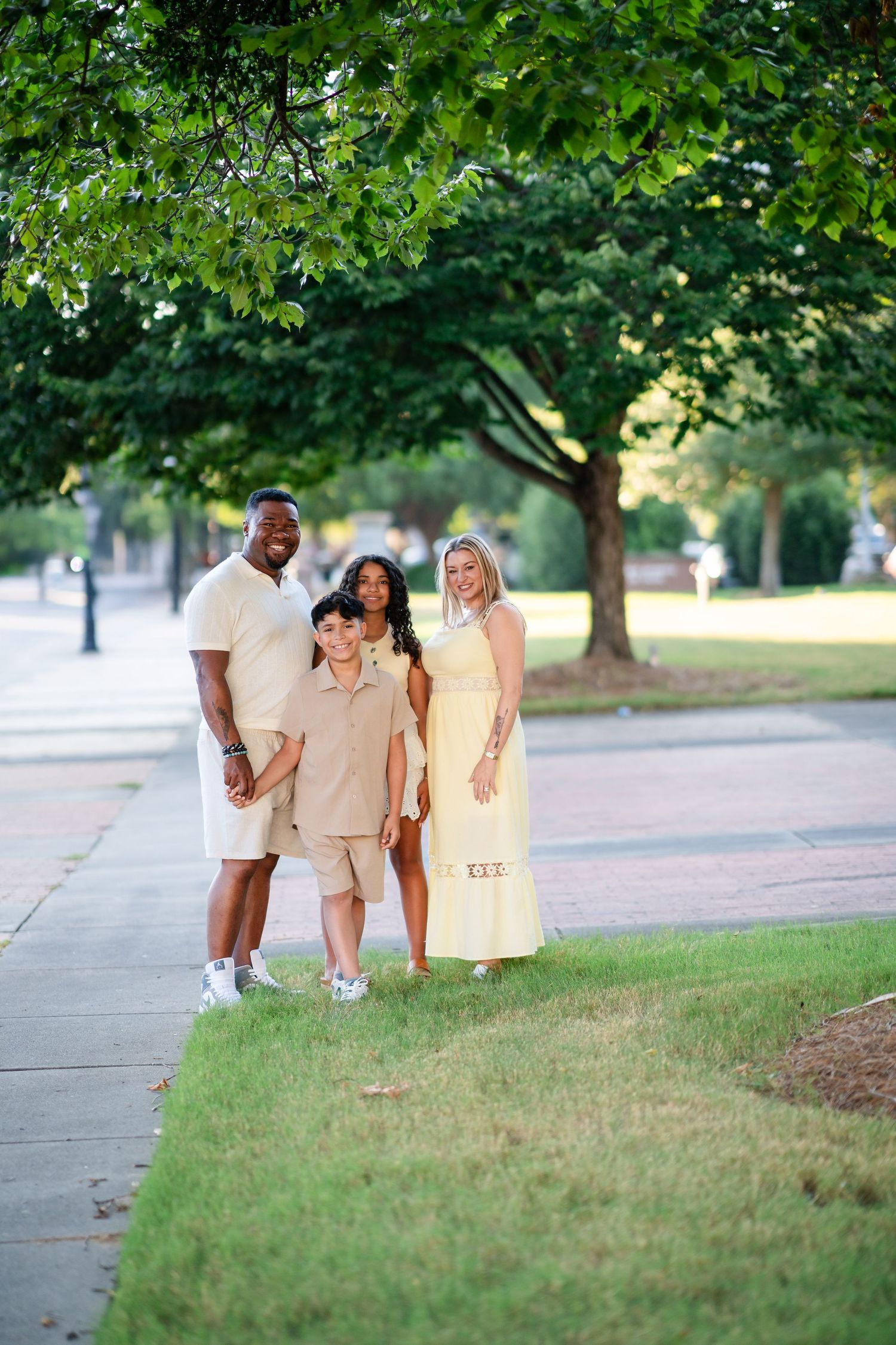 A joyful interracial family of four poses outdoors on a sunny day, dressed in coordinating beige and yellow outfits.
