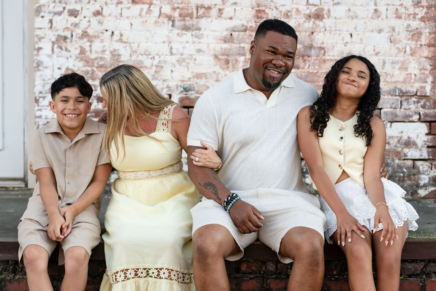 Happy family of four sitting together outdoors, smiling in coordinated light outfits against a brick wall.