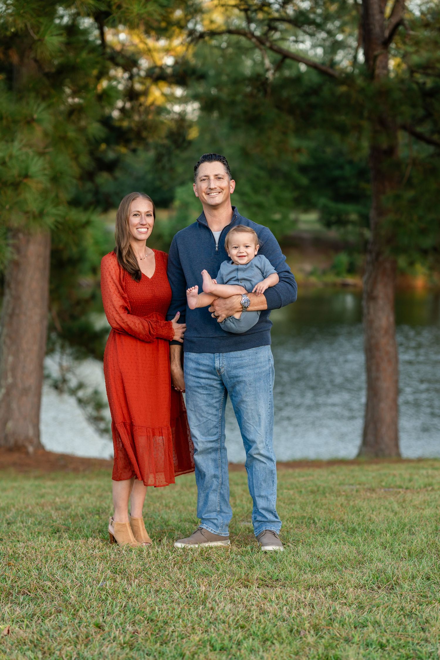 Family portrait of smiling couple with baby boy standing on green grass near a pond surrounded by trees.