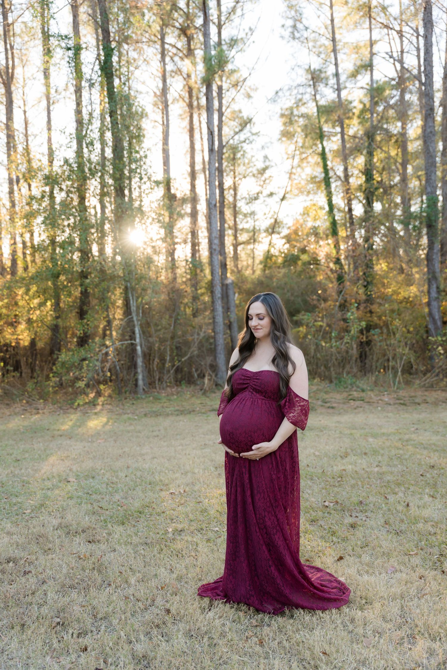 A maternity portrait showing an expectant mother in a flowing burgundy dress amid tall autumn trees.