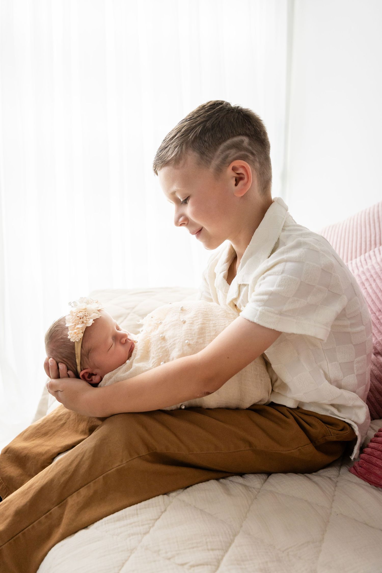 Sweet interaction between siblings lying on white bedding with natural window light.