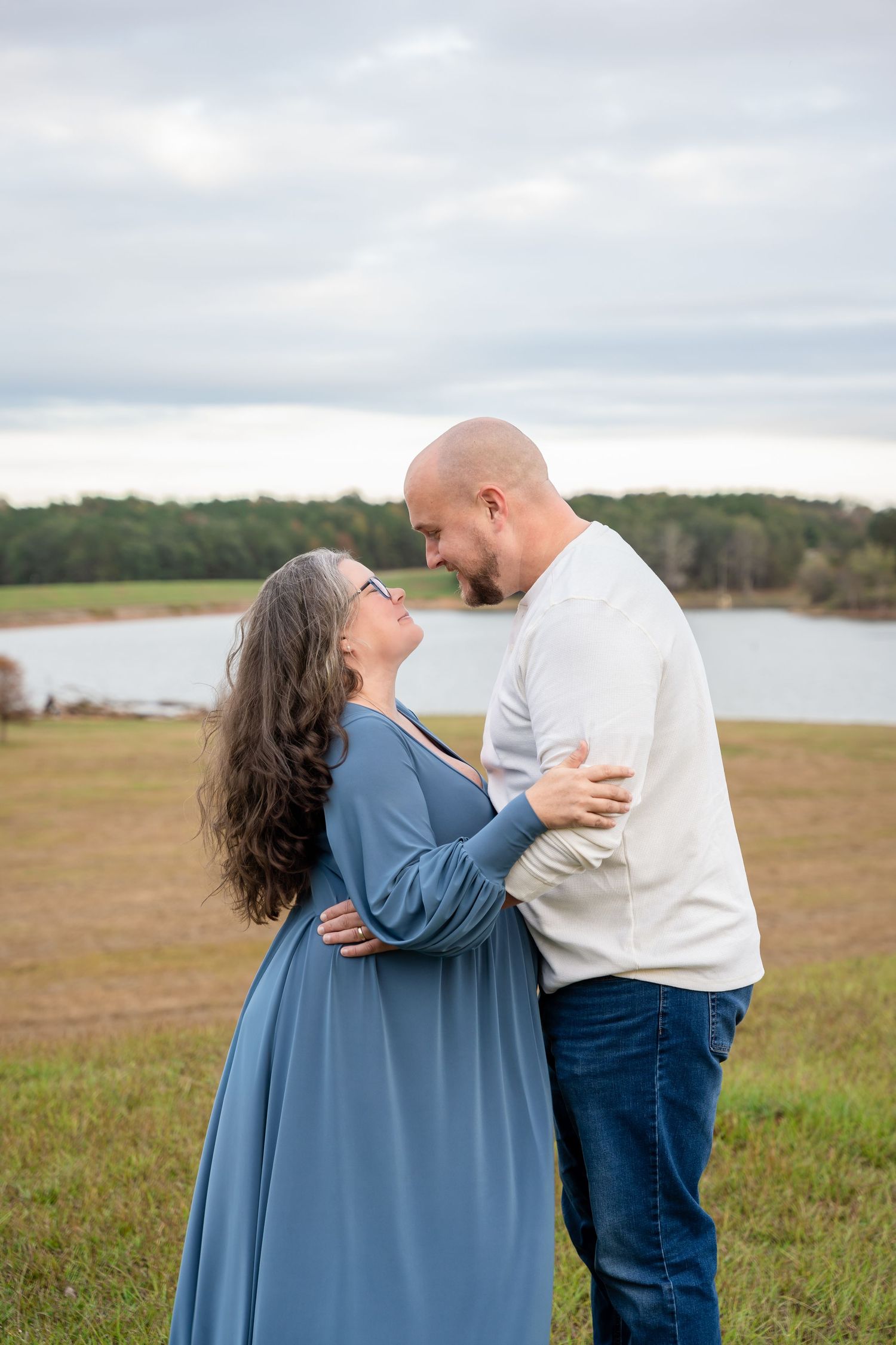 Romantic outdoor photoshoot on grassy field with lake view and overcast sky.