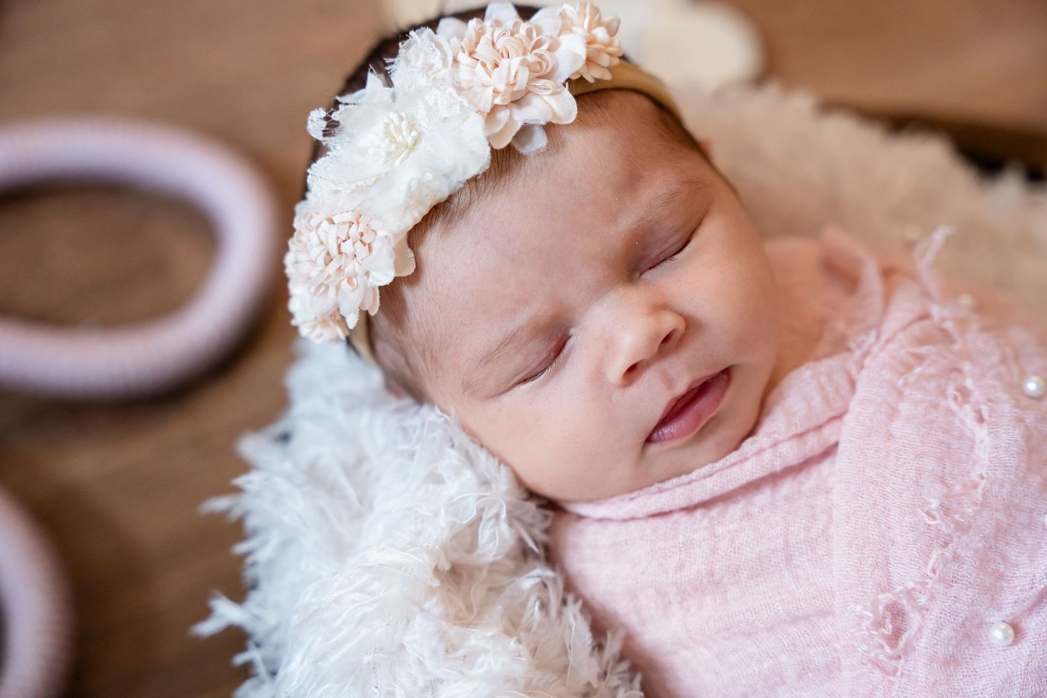 Sleeping newborn baby girl wrapped in pink swaddle with floral headband on fluffy white blanket.