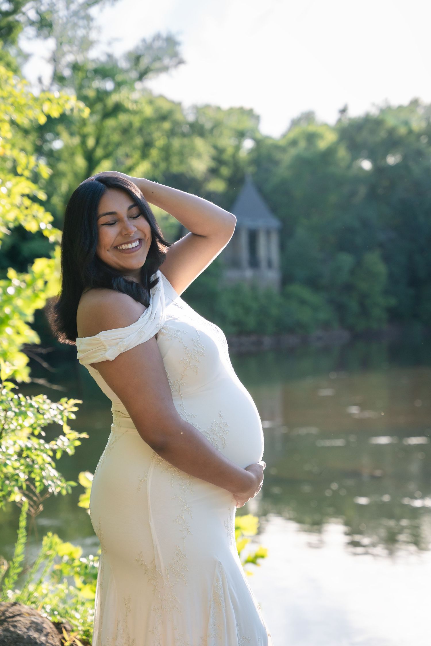 Smiling pregnant woman in white off-shoulder gown cradling her belly outdoors near a serene pond with lush greenery.