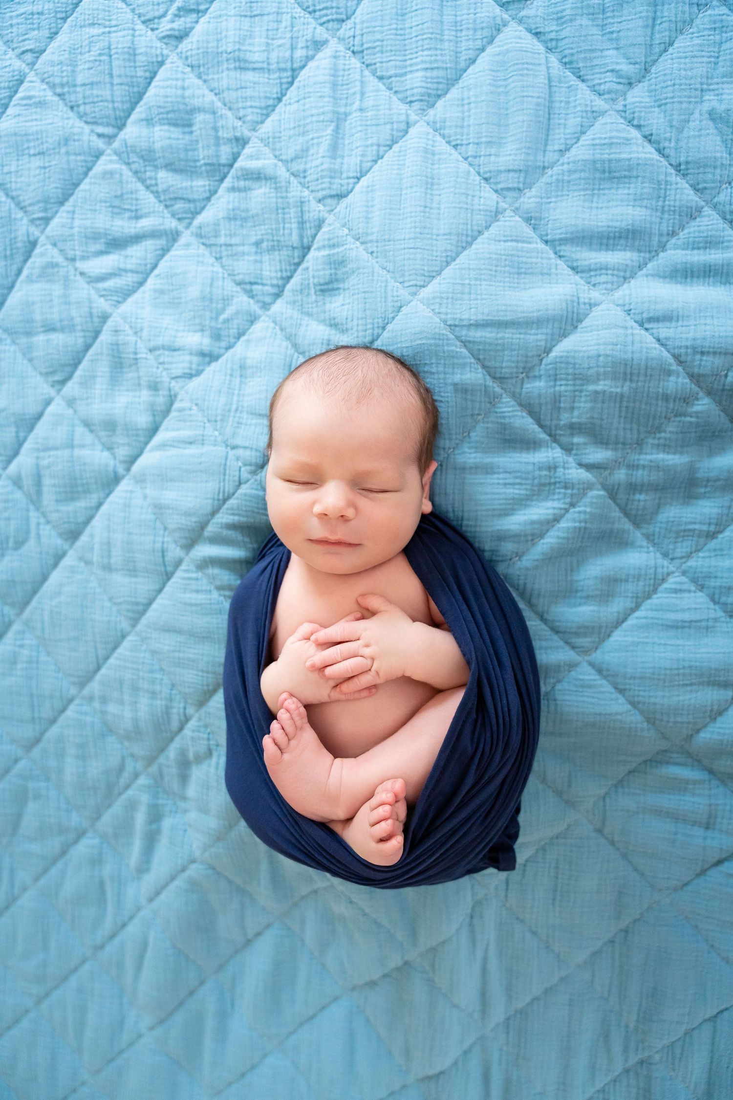 Sleeping newborn swaddled in dark blue fabric on quilted light blue background.