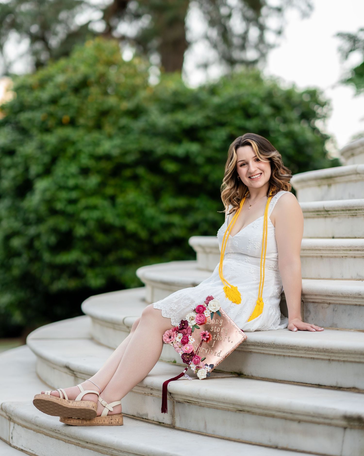 Graduate poses elegantly on white stone steps with yellow tassel and floral bouquet.