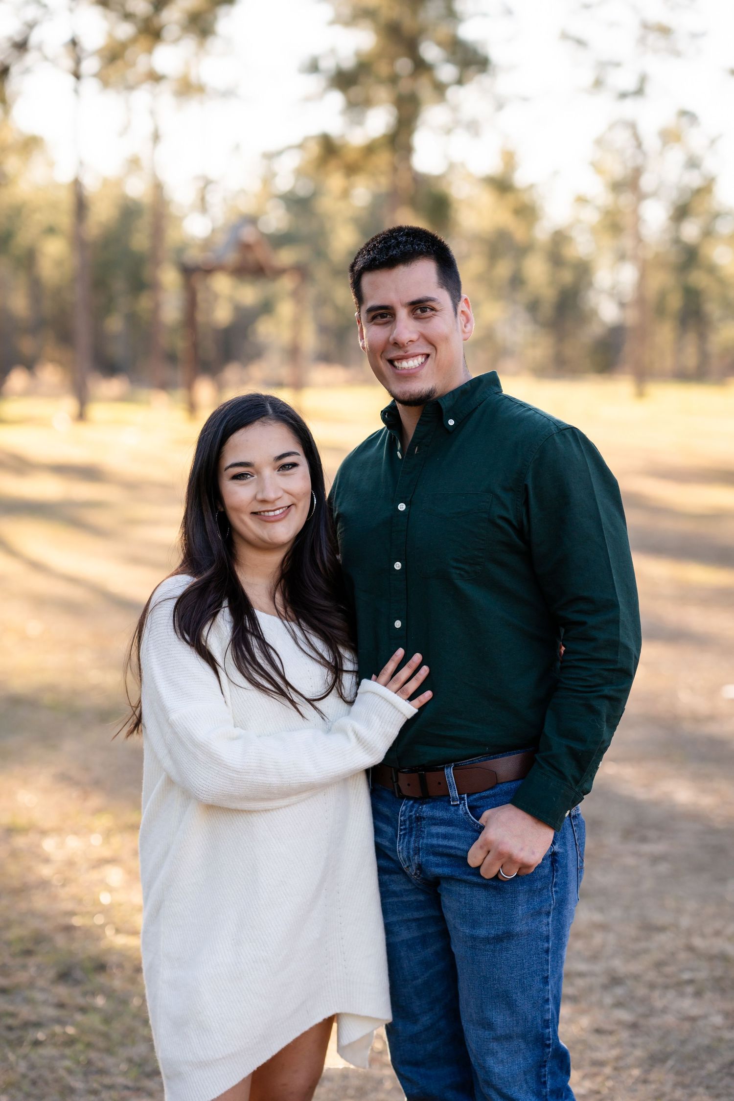 A couple in casual winter attire smiles at the camera in an outdoor forest setting.