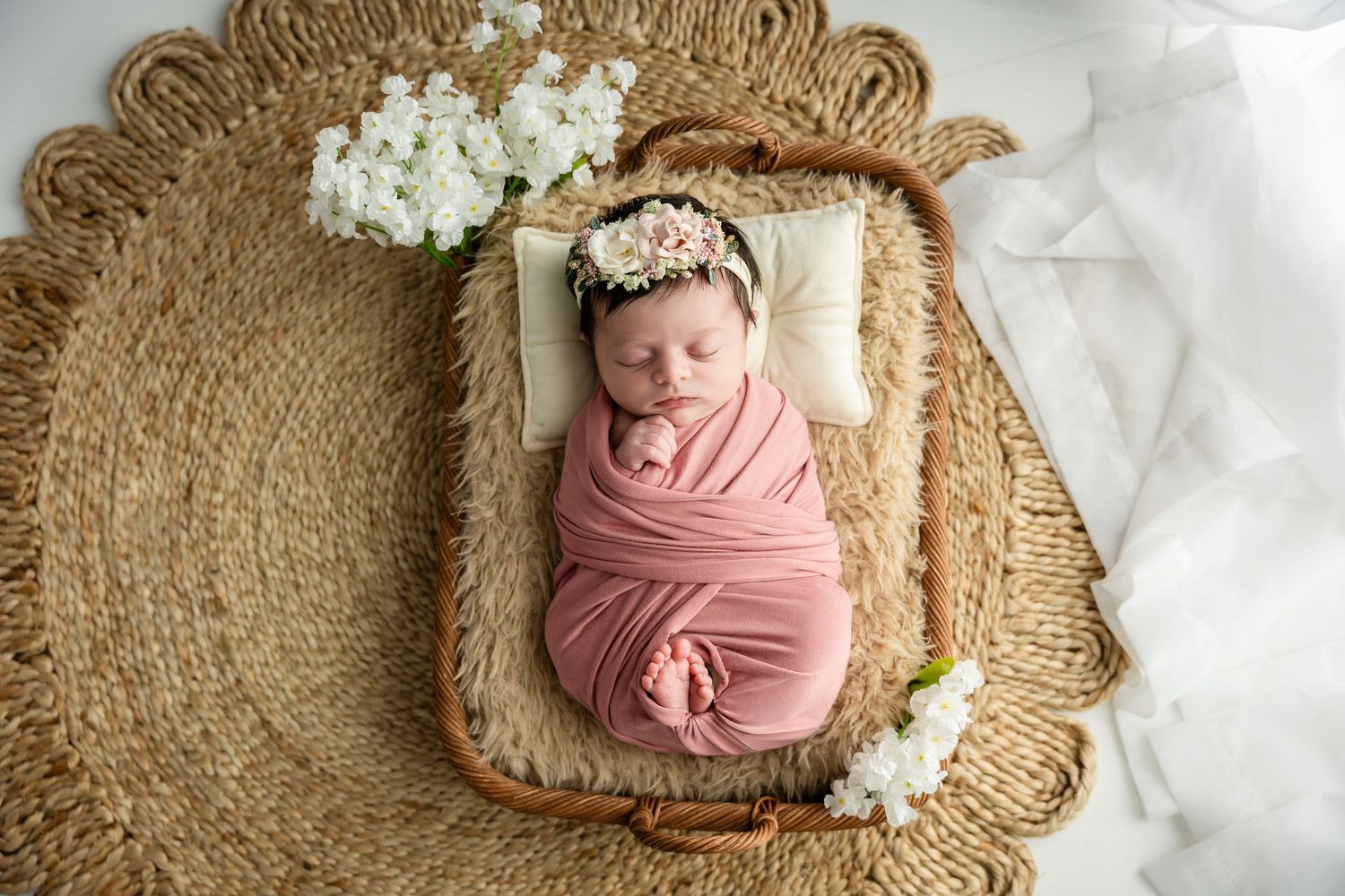 A newborn wrapped in soft pink fabric rests in a woven basket decorated with delicate white flowers.