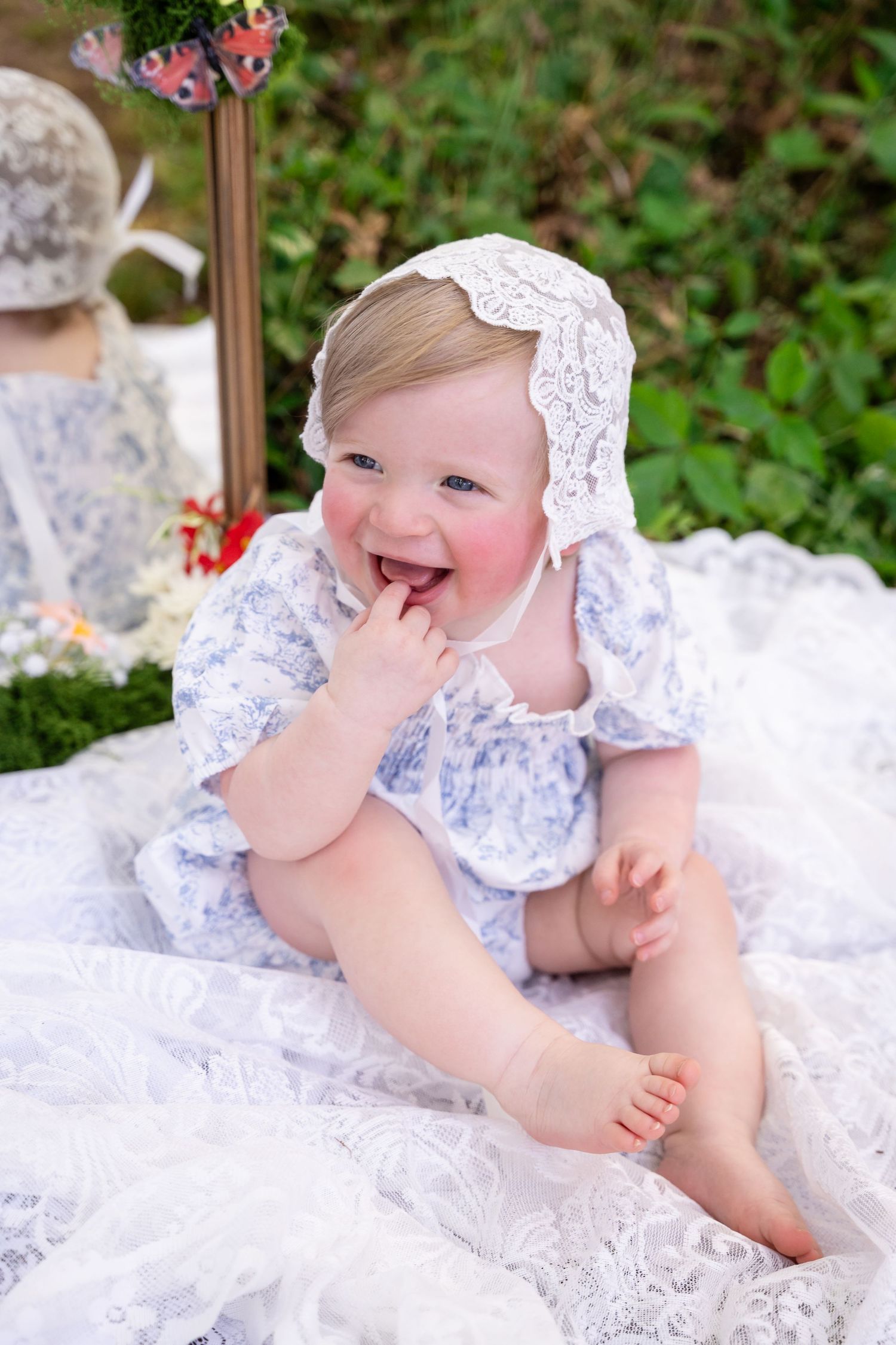 Happy baby girl in blue floral romper and lace bonnet sitting on white lace blanket outdoors.