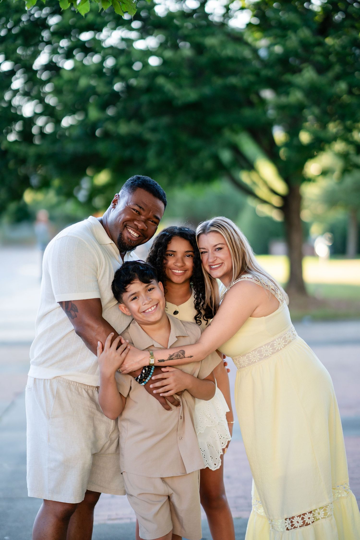 Happy interracial family of four hugging outdoors with lush green trees in the background.