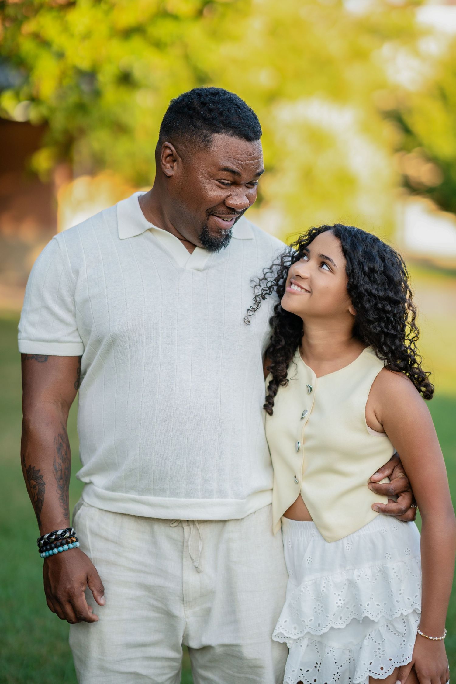 A smiling man and young woman in white outfits share a warm embrace outdoors in a sunny green park.