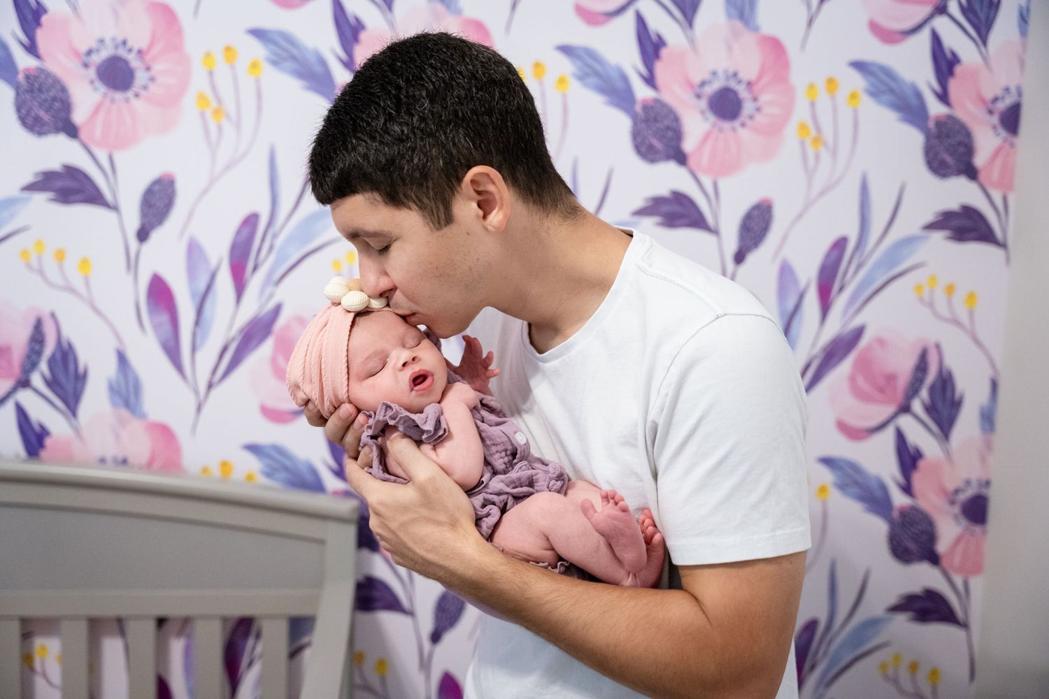 A father tenderly kisses his sleeping newborn baby girl dressed in pink in a floral nursery.