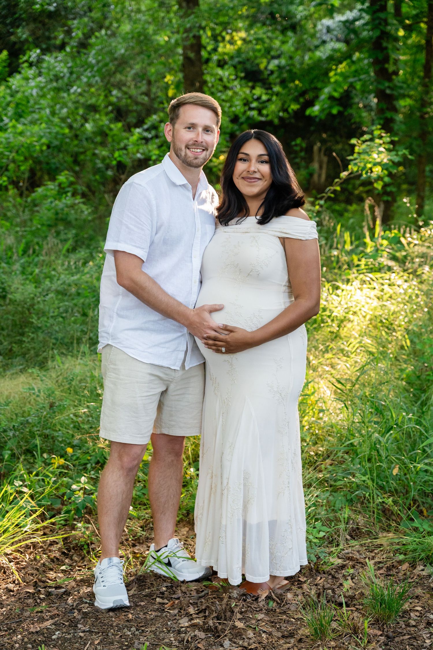Couple in white outfits posing outdoors, woman in white maternity dress cradling baby bump, surrounded by lush greenery.