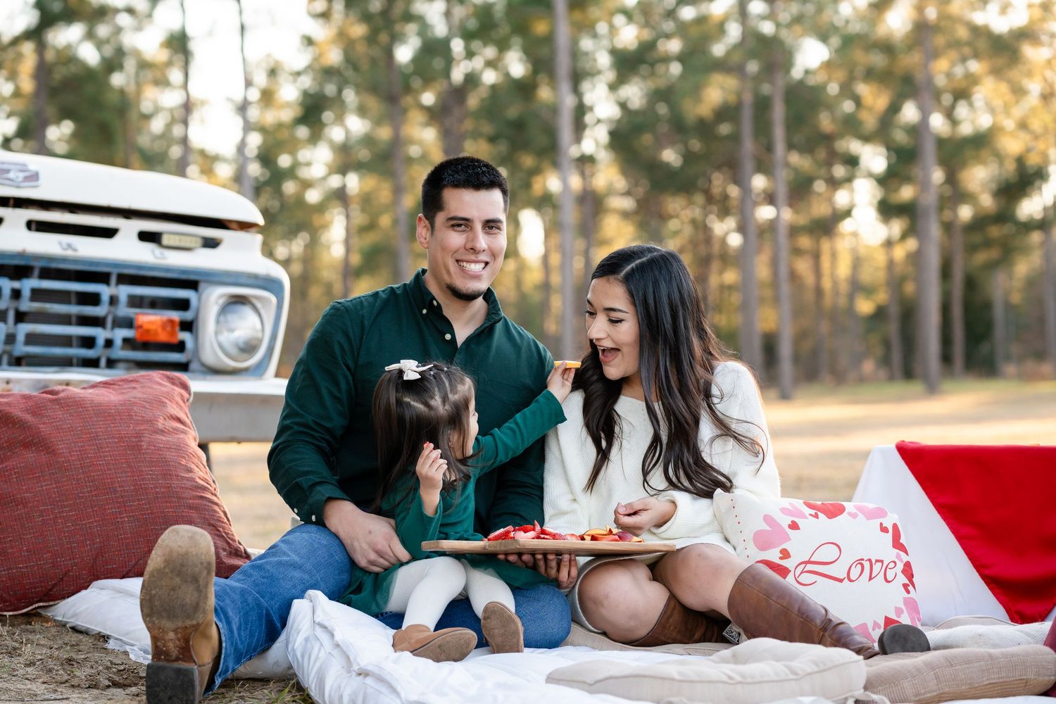 Family enjoying a picnic on blankets beside vintage truck in wooded setting.