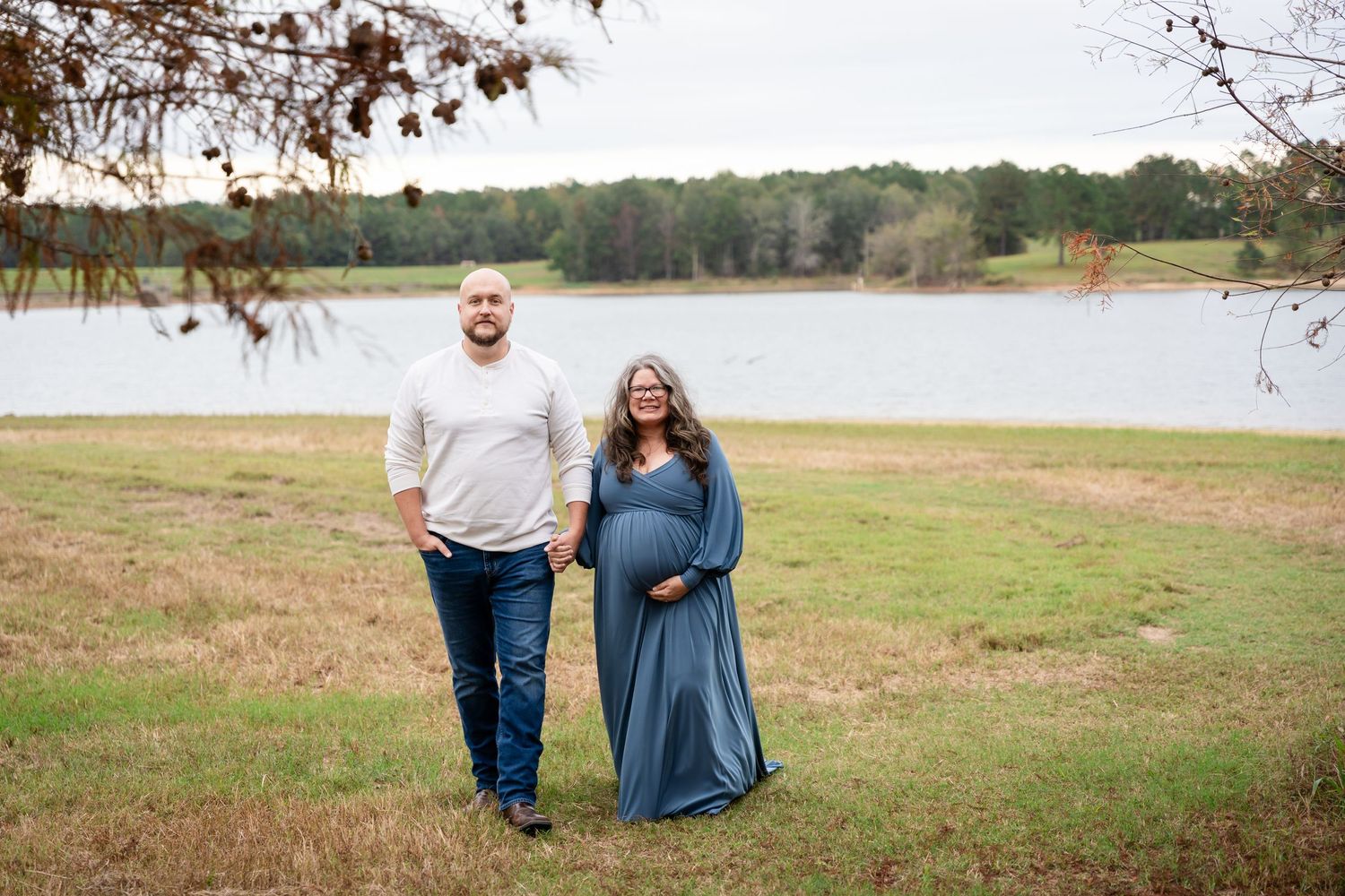 Romantic couple poses together on grassy field with lake and trees in background.