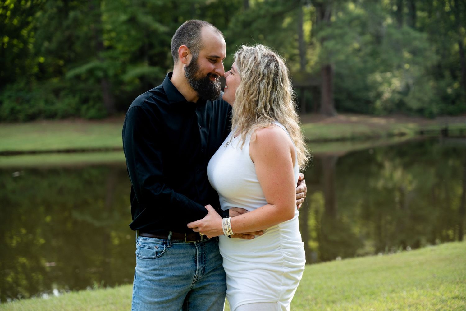 Couple sharing romantic moment on grassy lawn with lake and trees behind them.