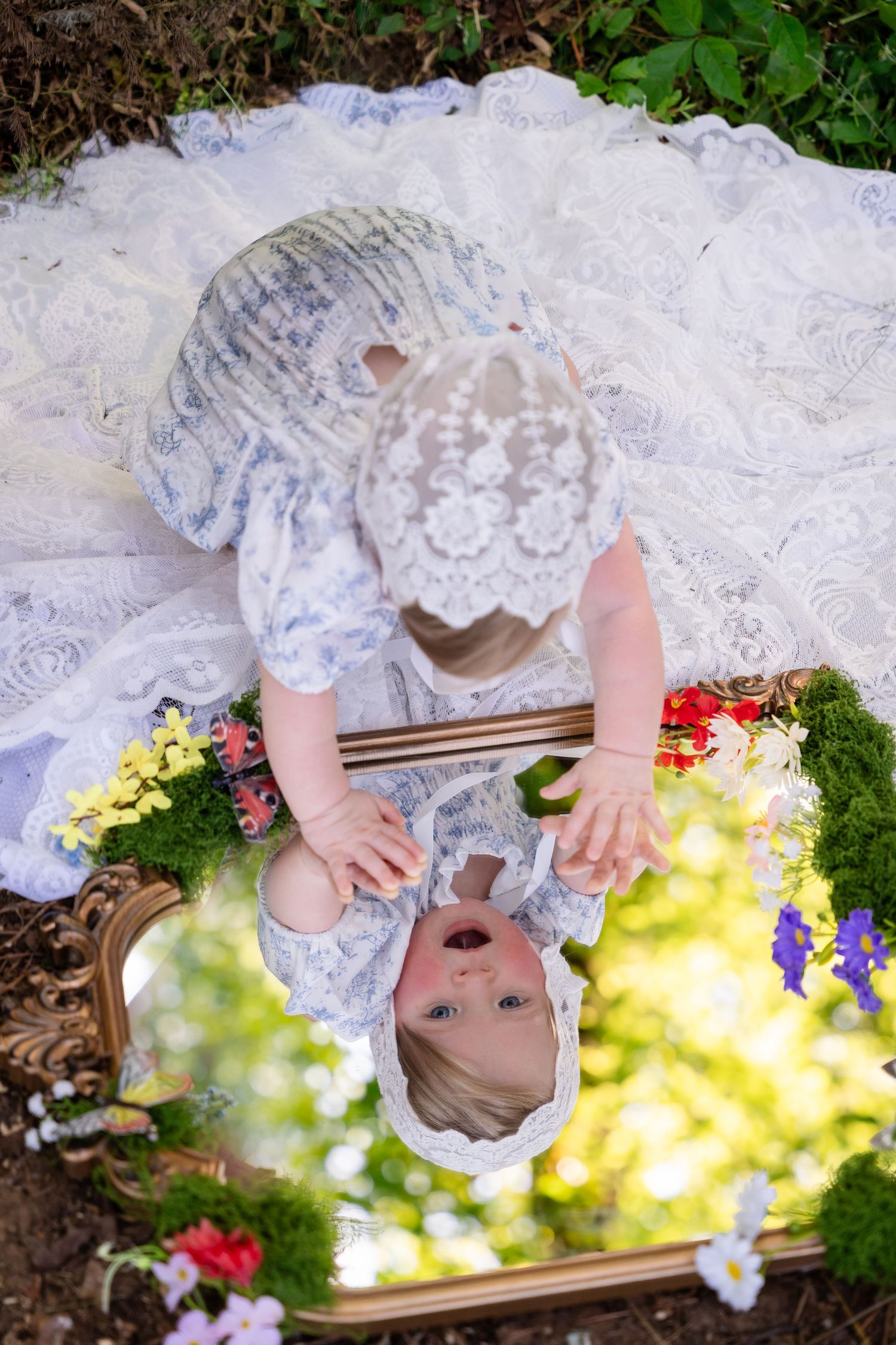 Aerial view of a baby in a white lace bonnet and dress looking into a gold-framed mirror surrounded by colorful flowers.