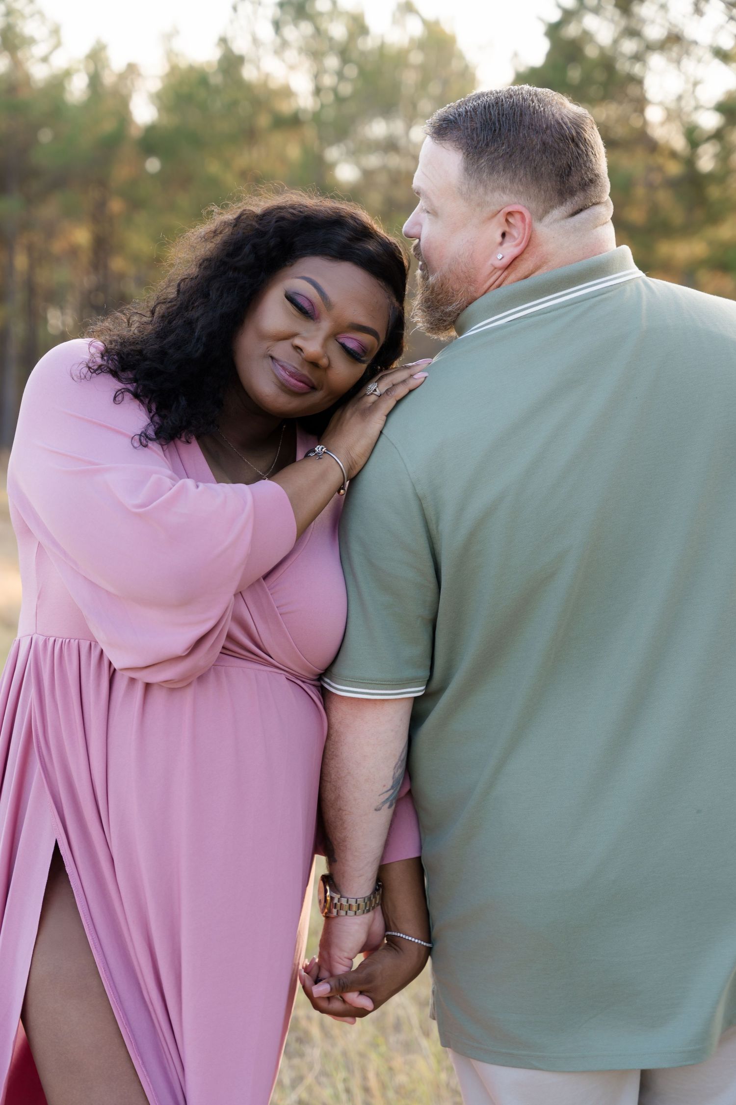 Interracial couple holding hands outdoors, woman in pink dress smiling, man in green polo shirt, forest background.