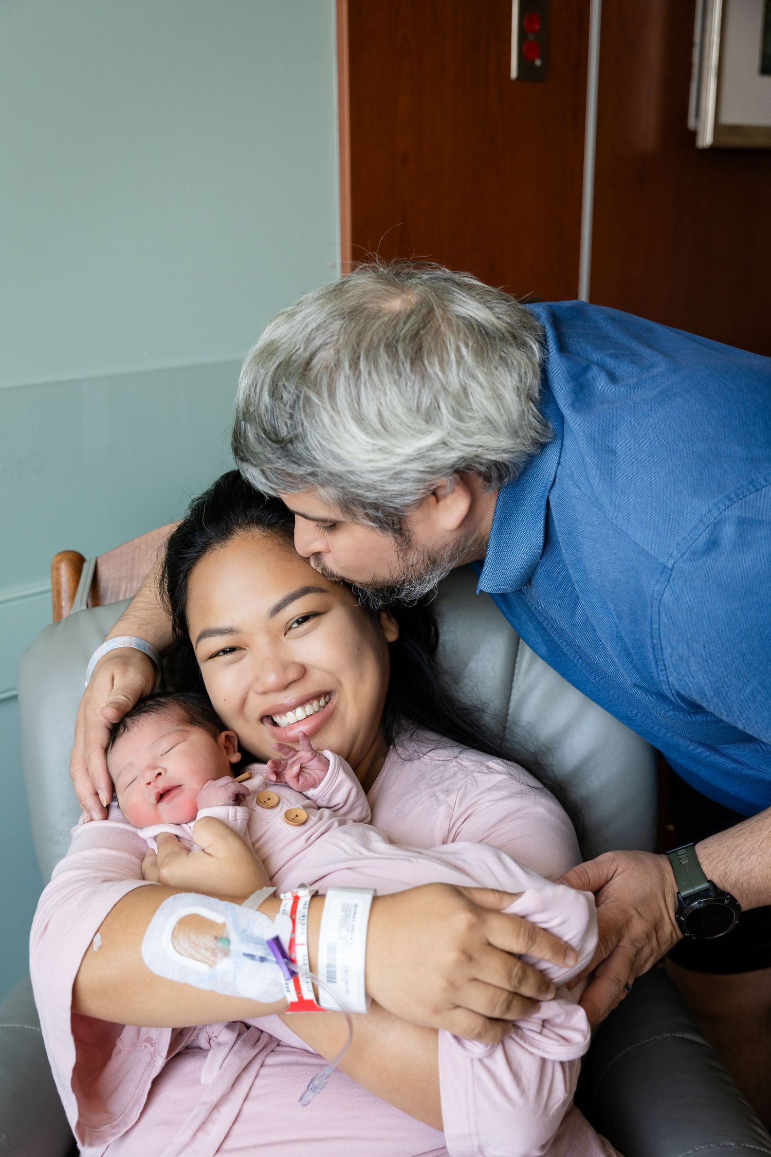 Smiling new mother in a hospital chair cradles her sleeping newborn baby while her gray-haired husband leans down to kiss her on the head.