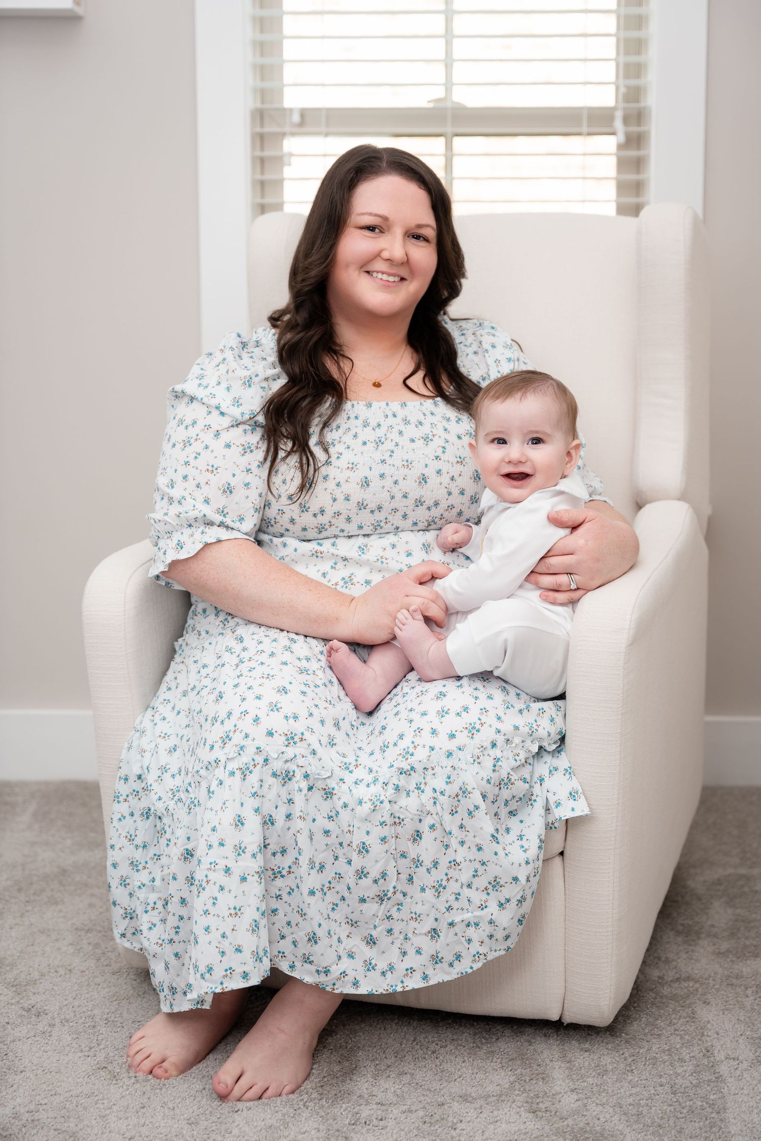 A floral dress-clad woman sitting in a beige armchair holding a baby in white.