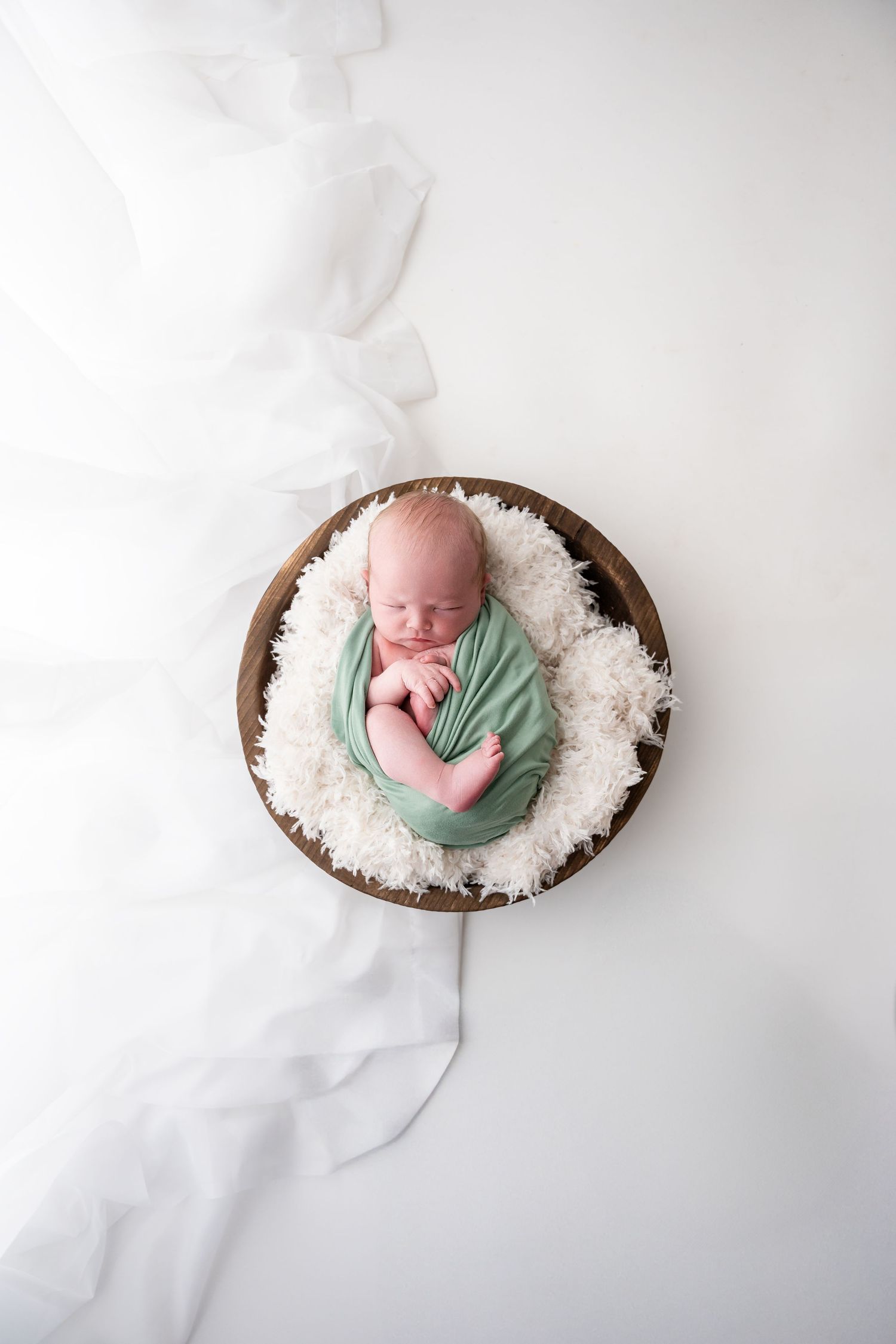 Sleeping newborn cradled in white textured bedding inside a wooden bowl on white background.