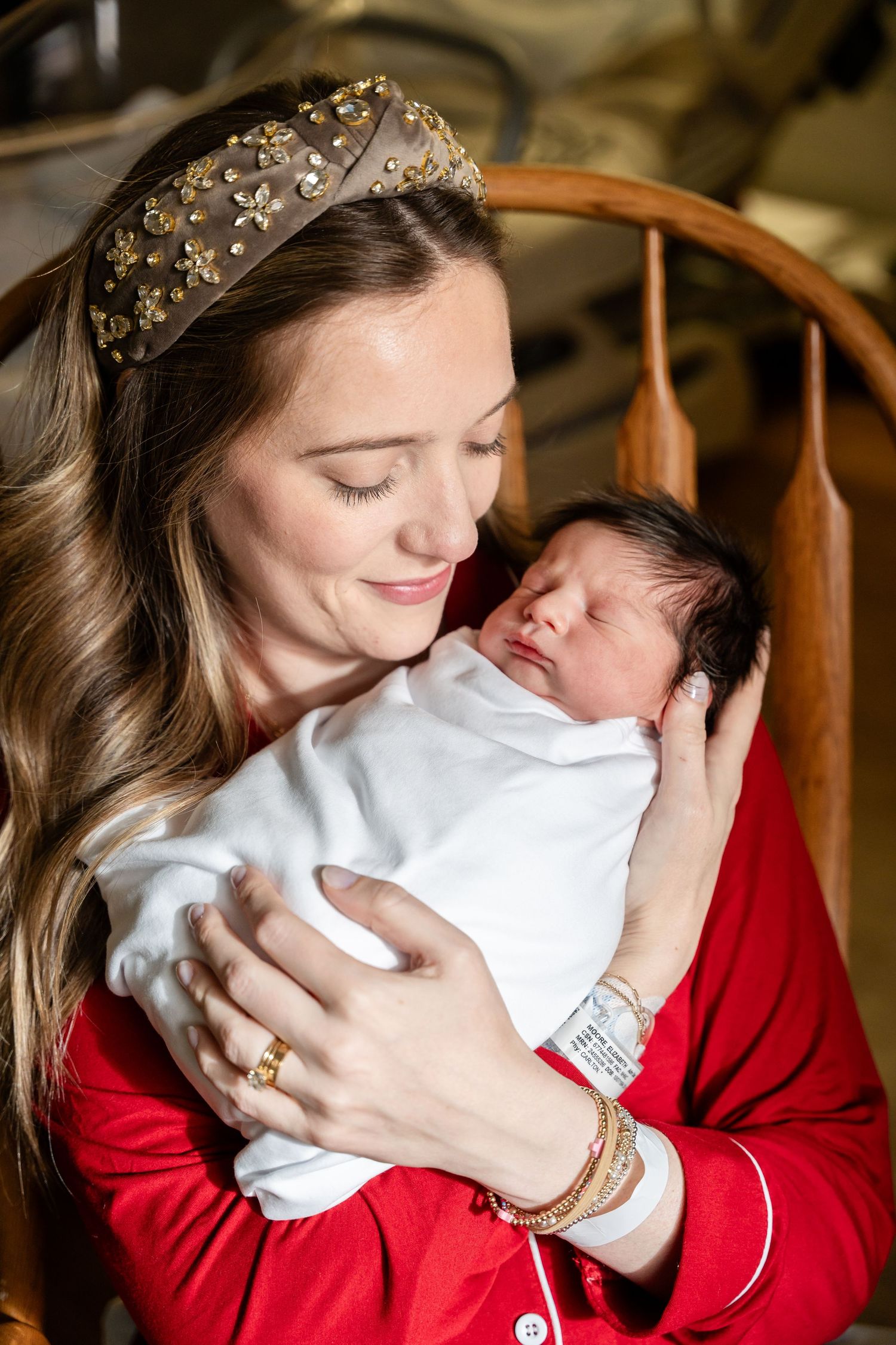 Woman in red sweater and sparkly headband holding newborn wrapped in white blanket.