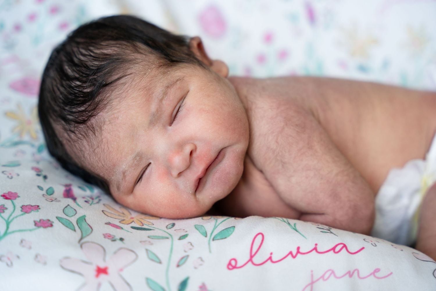 Sleeping newborn baby with dark hair lies on a floral blanket with the name Olivia Jane written in pink script.