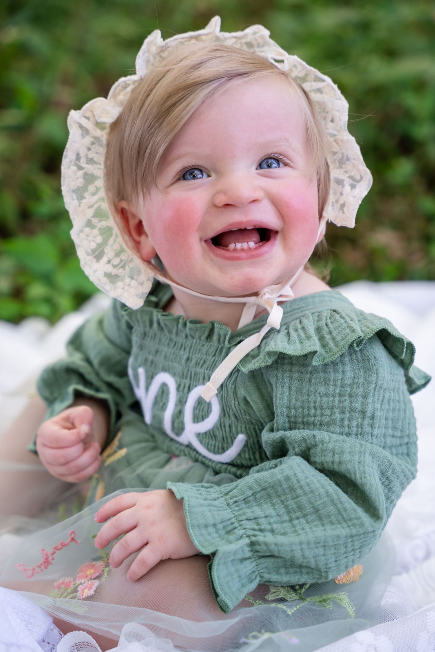 Smiling baby girl in green dress with 'one' embroidery and lace bonnet celebrating first birthday outdoors.