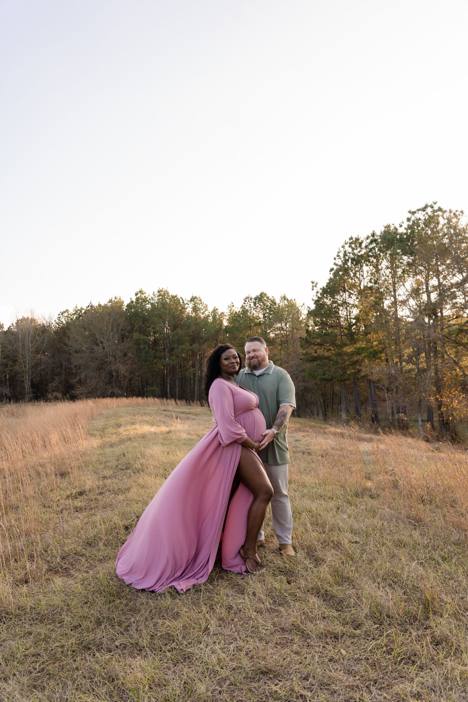 Pregnant couple posing in a golden field at sunset, woman in flowing pink maternity gown, trees in background.