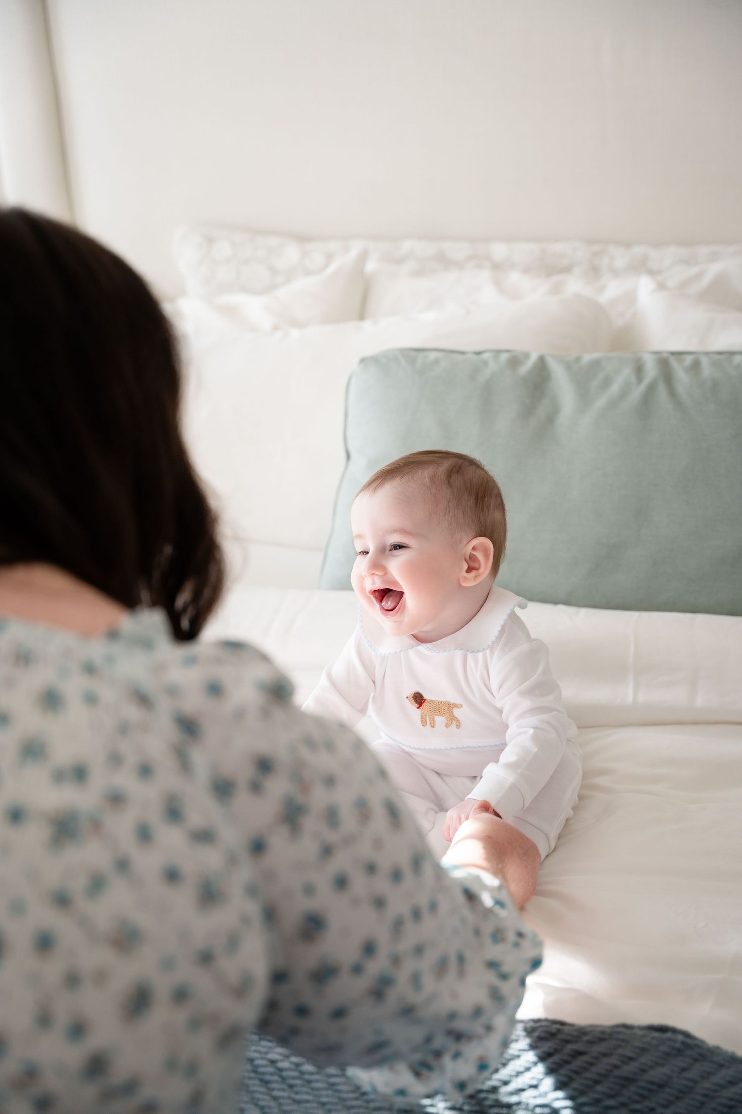Happy baby in white onesie playing on bed with sage green pillows.