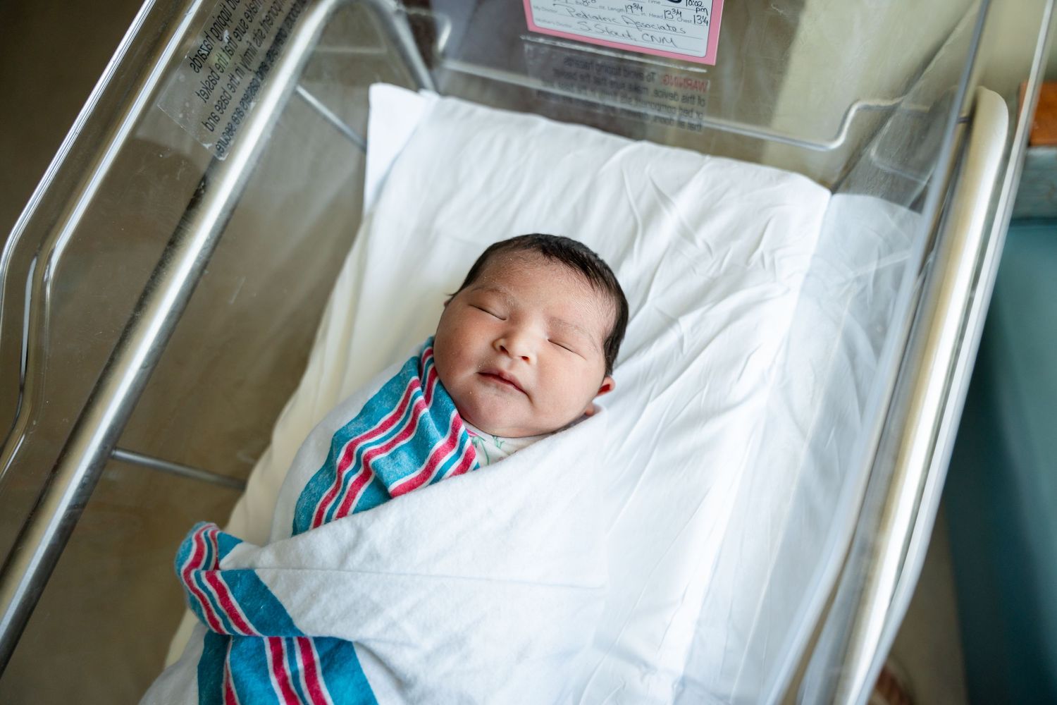 Sleeping newborn baby wrapped in a striped hospital blanket lies peacefully in a clear bassinet in a hospital maternity ward.