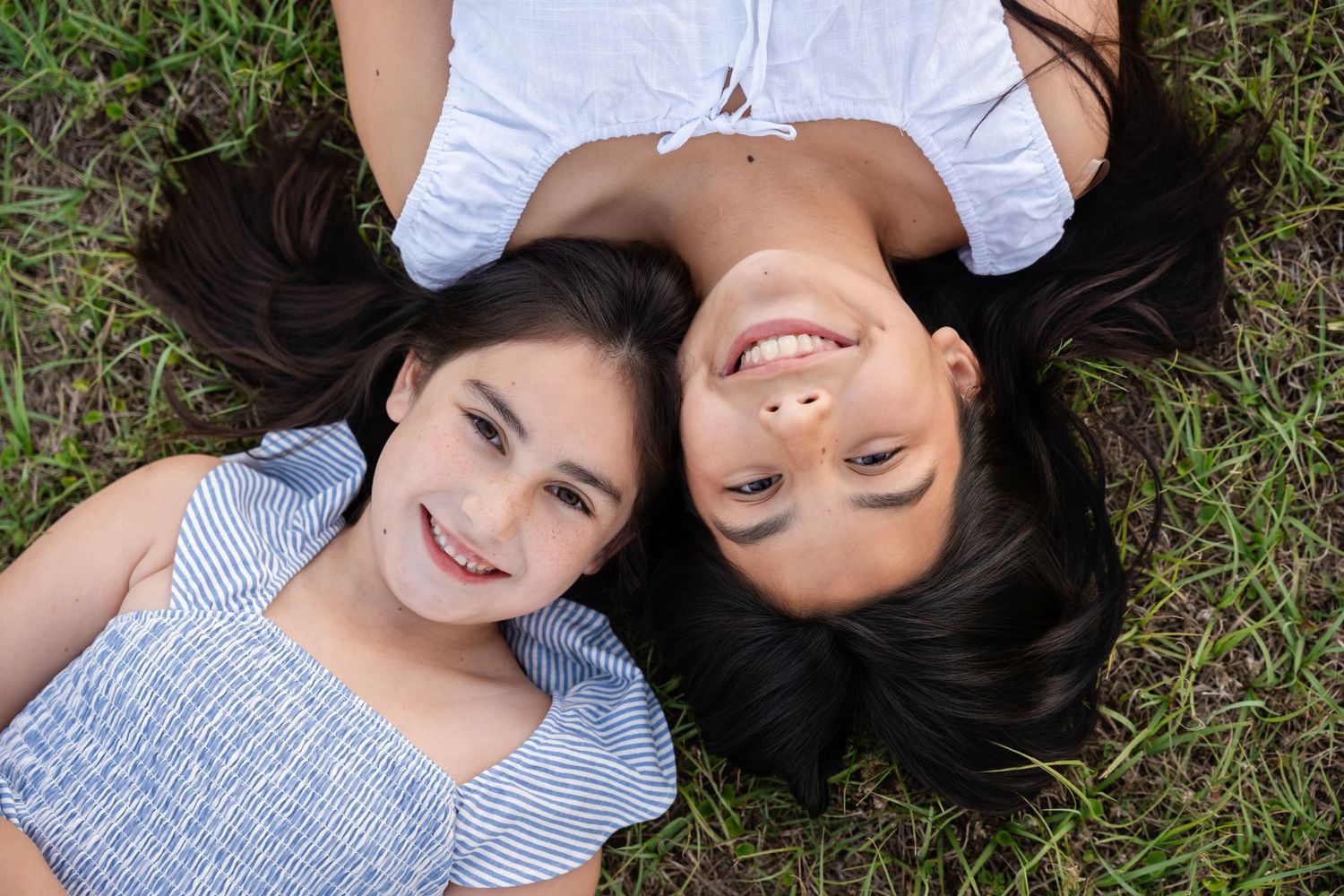 Two smiling girls with long dark hair lie on their backs in green grass, heads together, as seen from directly above.