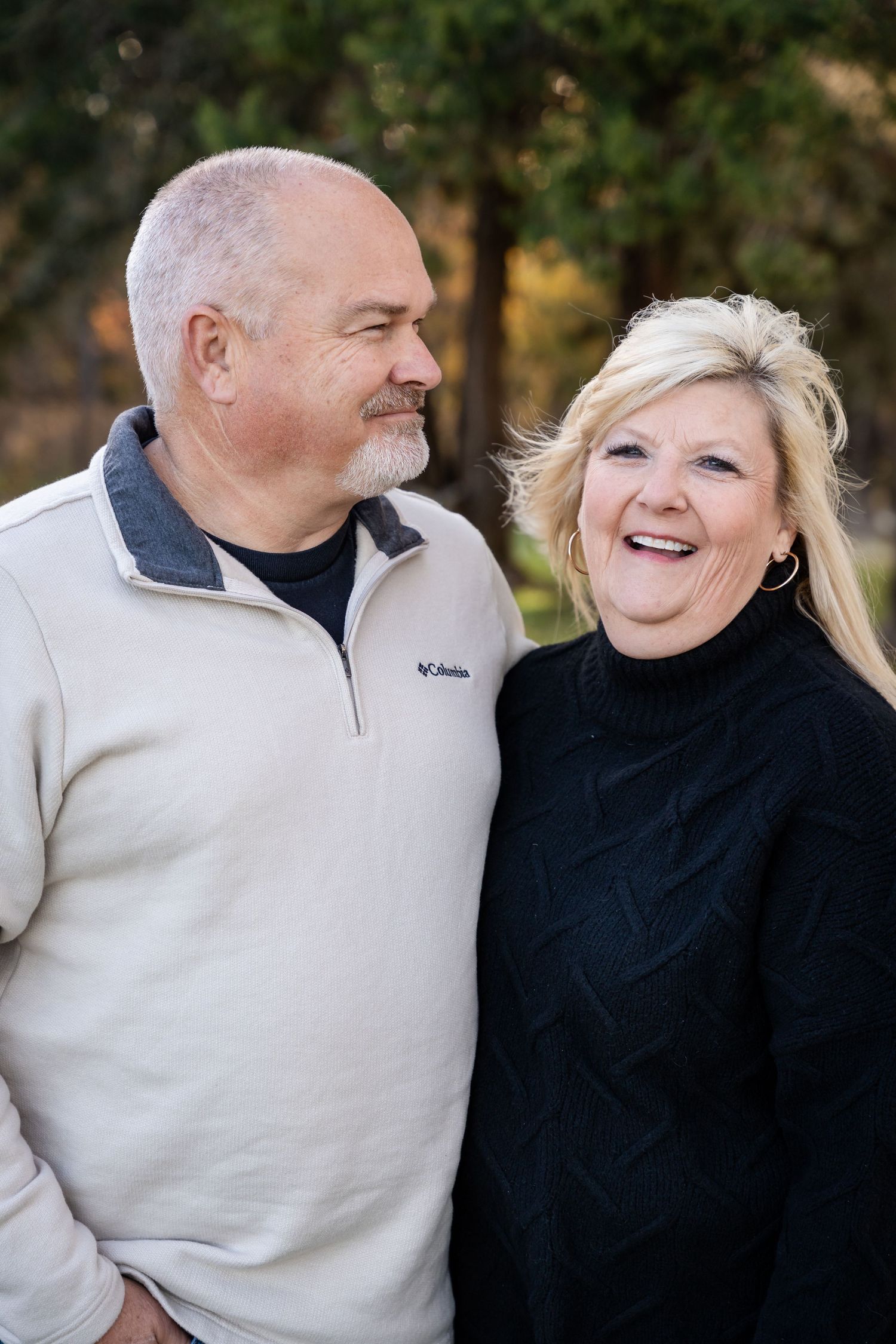 Smiling middle-aged couple standing close together outdoors, the man in a cream fleece pullover and the woman in a black sweater.