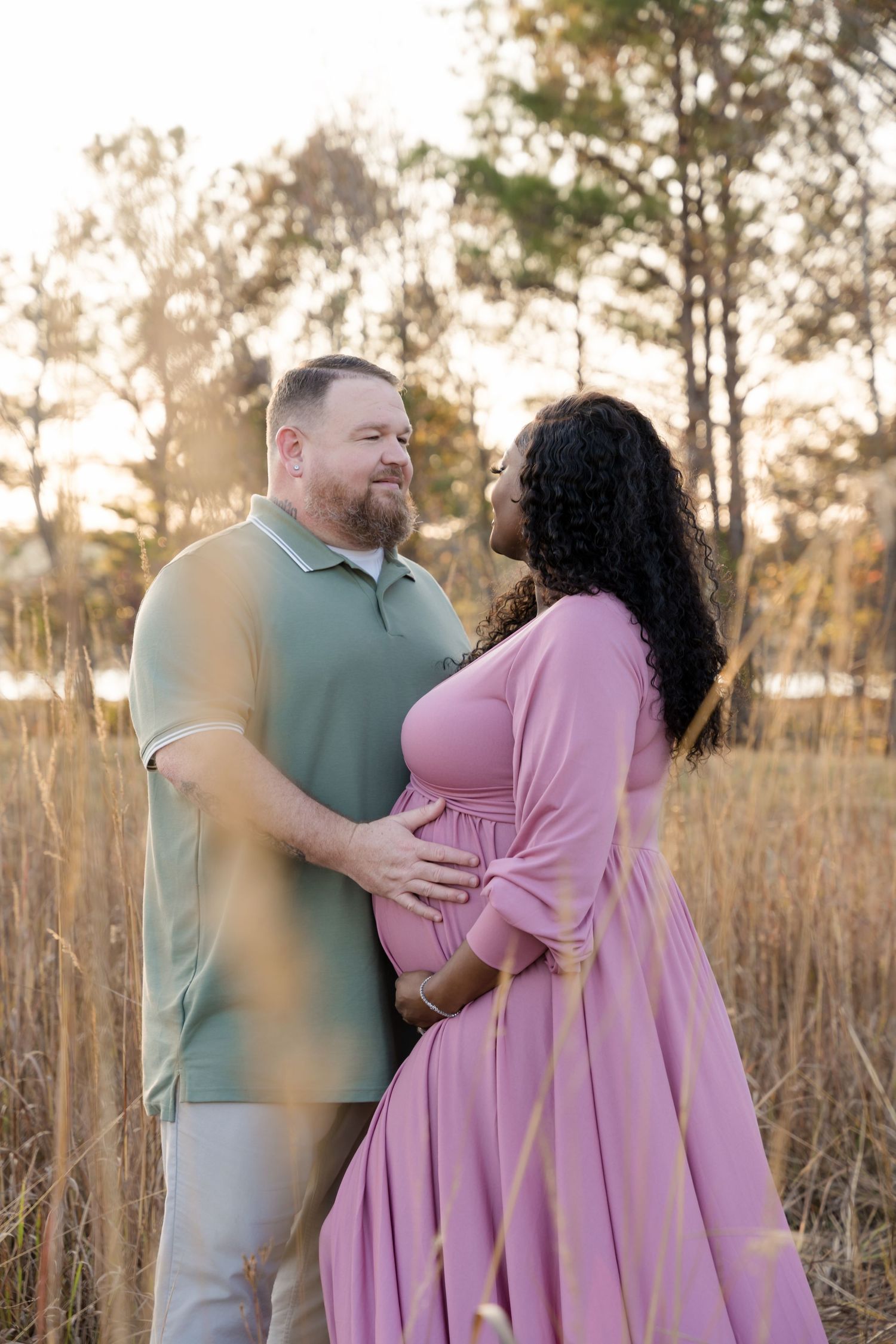 Couple sharing a tender moment in a golden field at sunset, woman in pink maternity dress cradling baby bump.
