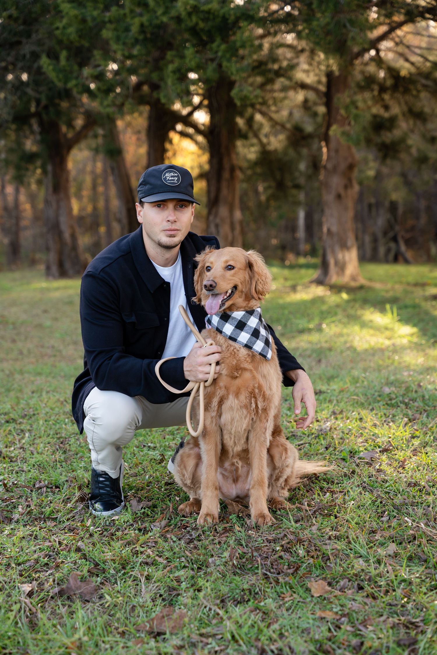 A man in a black cap and jacket crouches outdoors holding a leash beside a happy golden retriever wearing a plaid bandana.