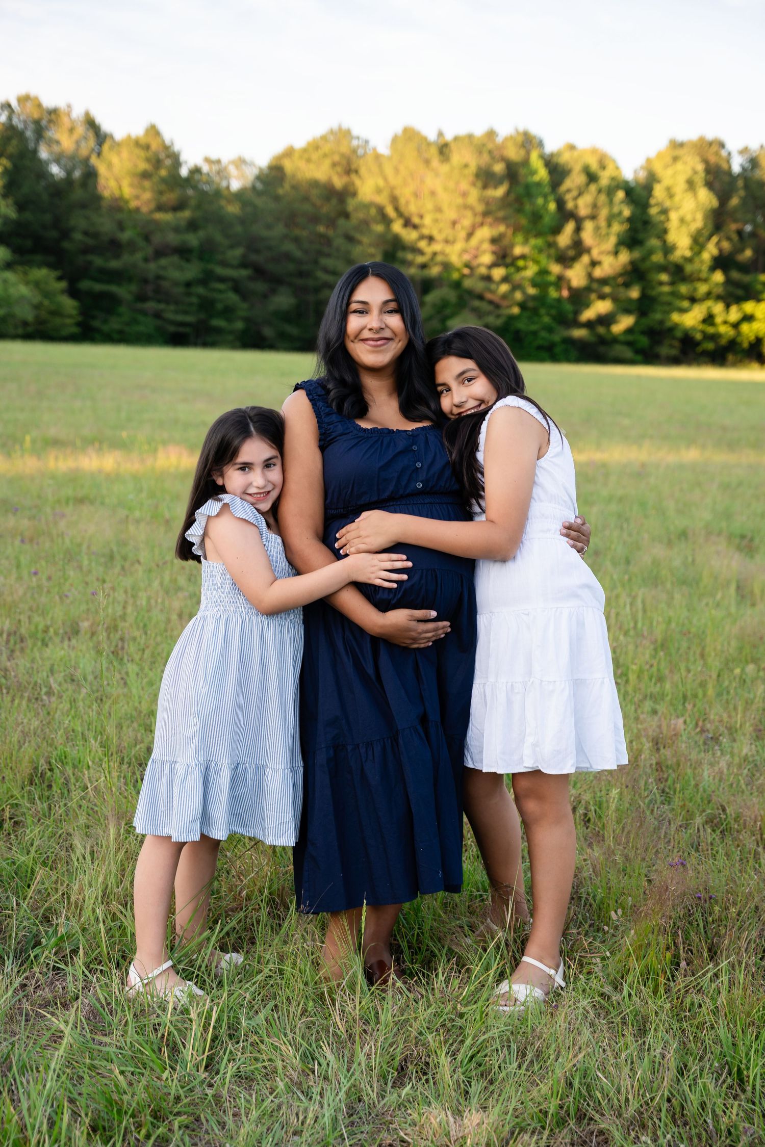 Expectant mother in a navy dress hugged by two smiling daughters in light dresses while standing in a grassy field at sunset.
