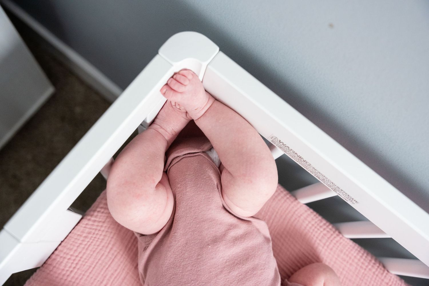Baby in pink onesie lying in a white crib with tiny feet resting on the crib rail.