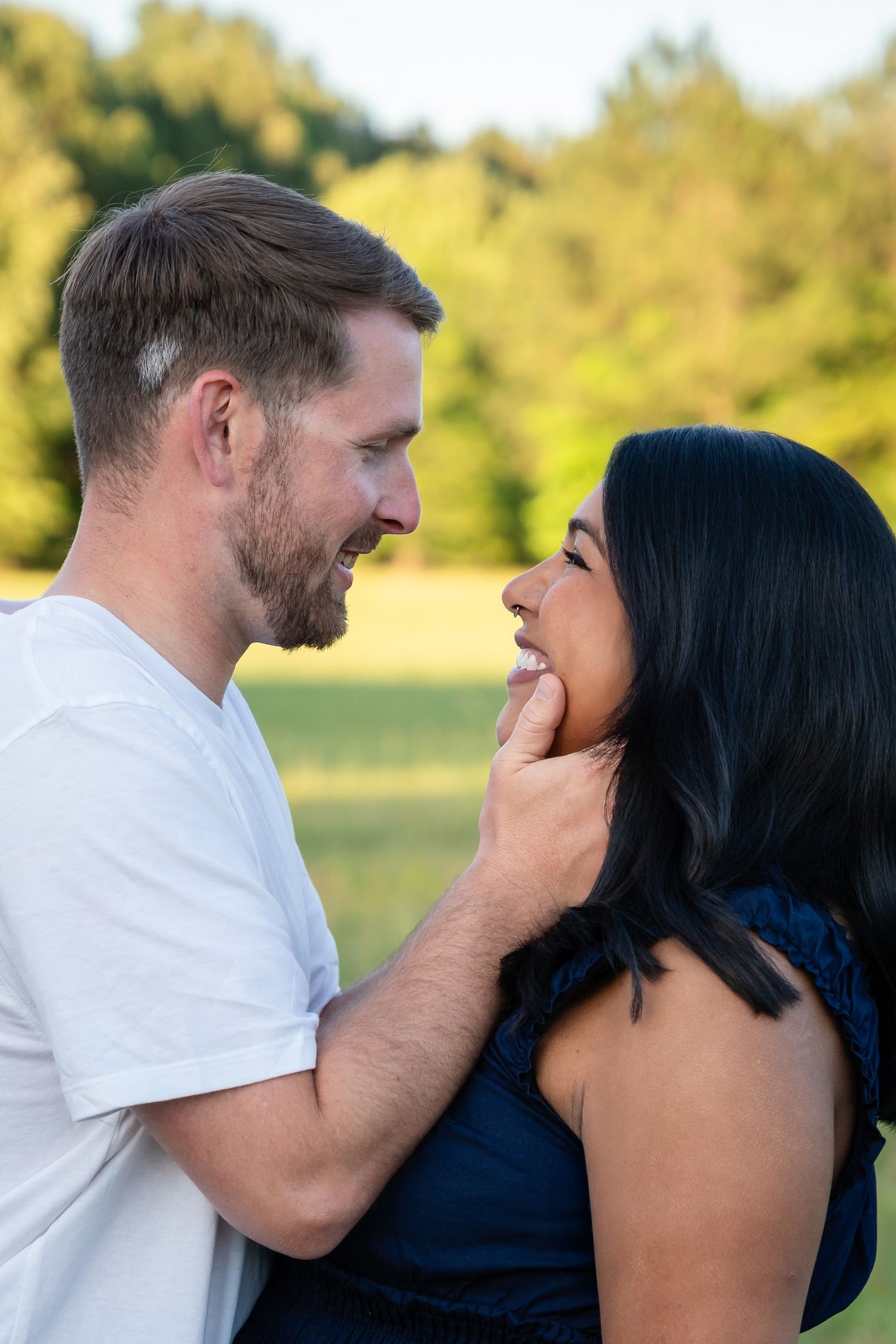 A smiling couple share a tender moment outdoors as a bearded man in a white shirt gently cups a woman's chin in a sunny green field.