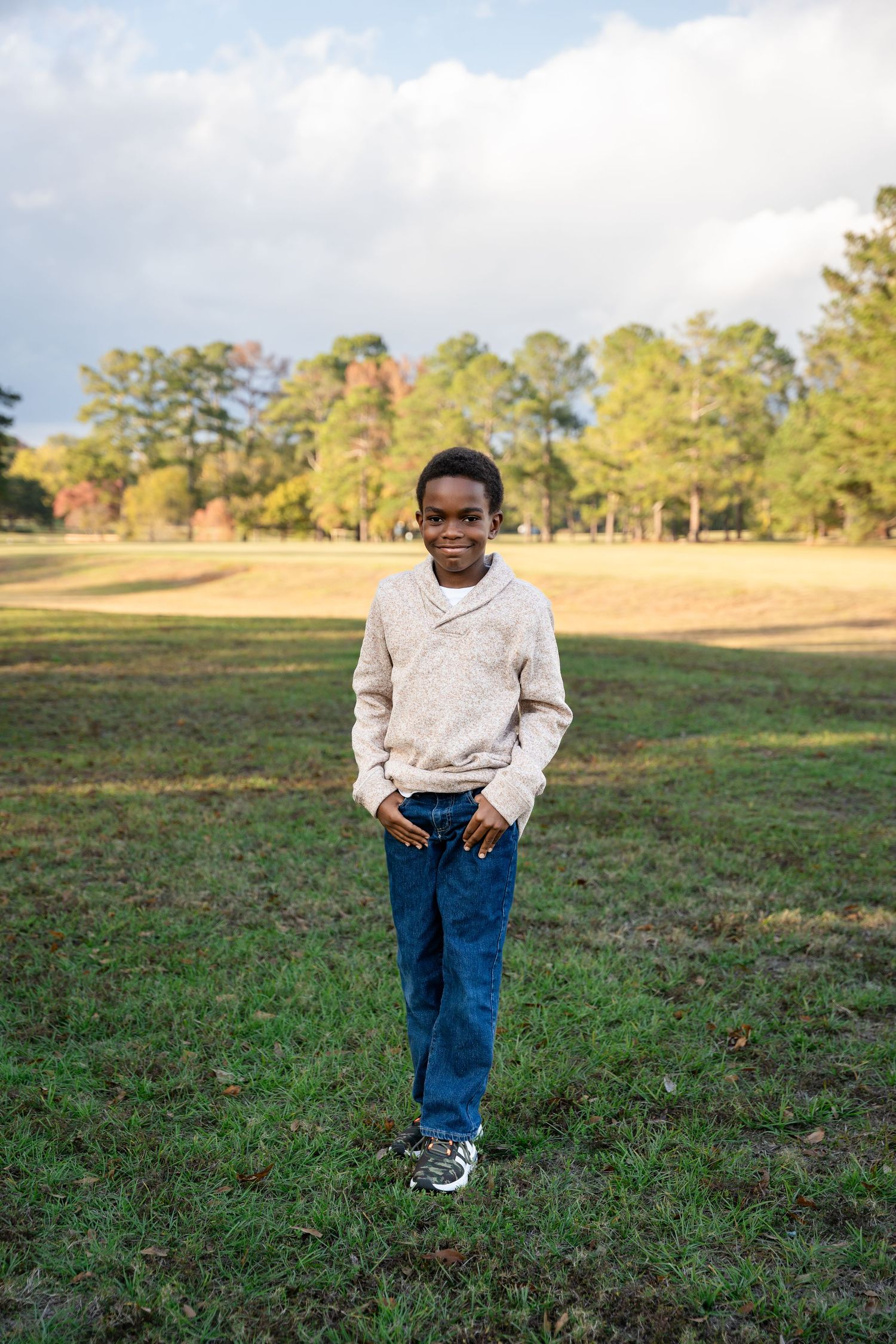 Person wearing white sweater and jeans stands in grassy field with autumn trees in background.