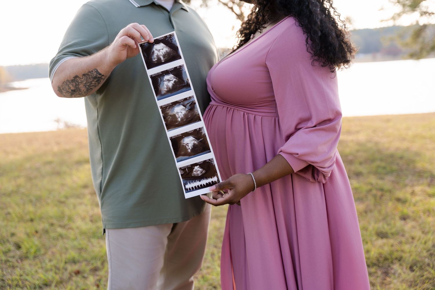 Pregnant couple holding ultrasound photos outdoors near a lake, woman in pink maternity dress, man in sage green shirt.