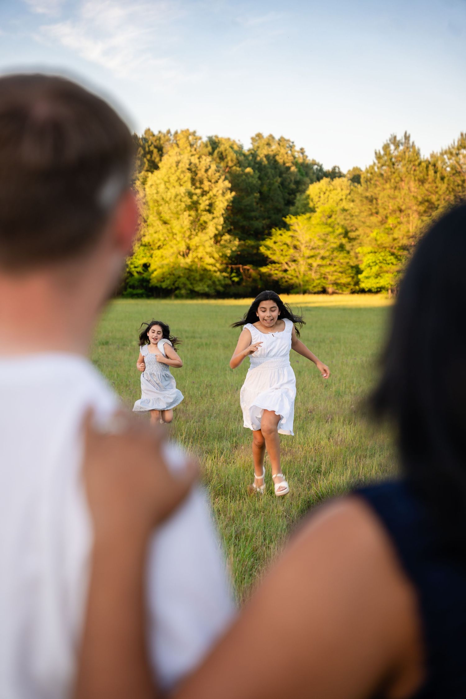 Two girls in white dresses running through a green field toward adults, with trees in the background on a sunny day.
