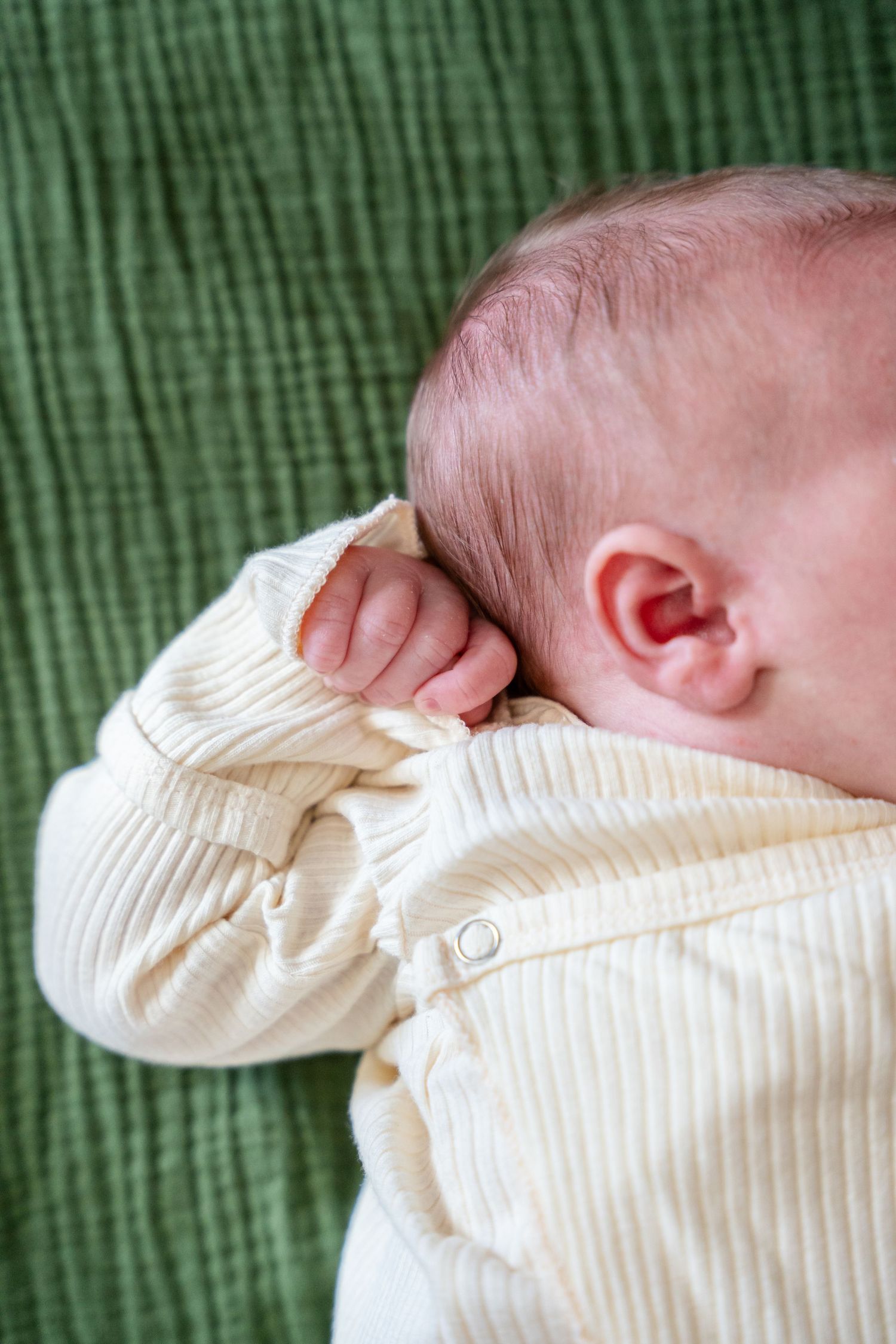 Newborn baby in cream ribbed onesie sleeping on green blanket, with tiny hand raised near face.