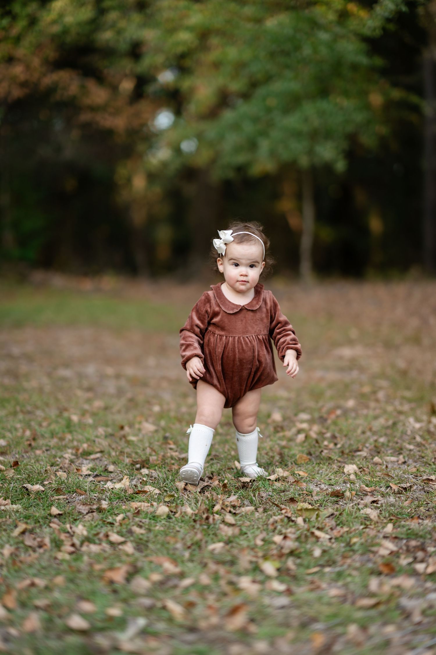 Small toddler in brown outfit stands alone on fallen autumn leaves.