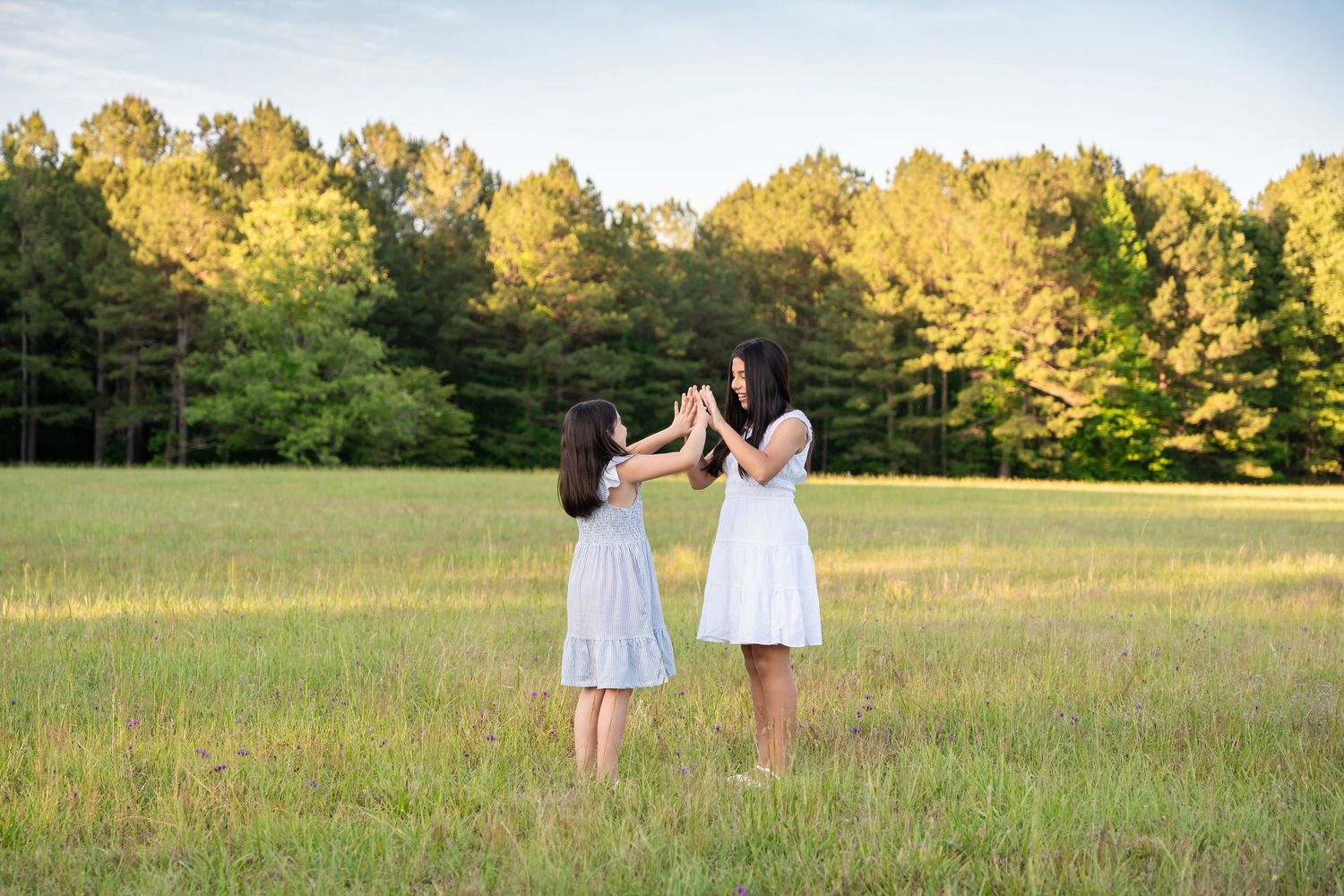 Two girls in white dresses stand facing each other with hands raised in a sunny grassy field, with tall green trees behind.