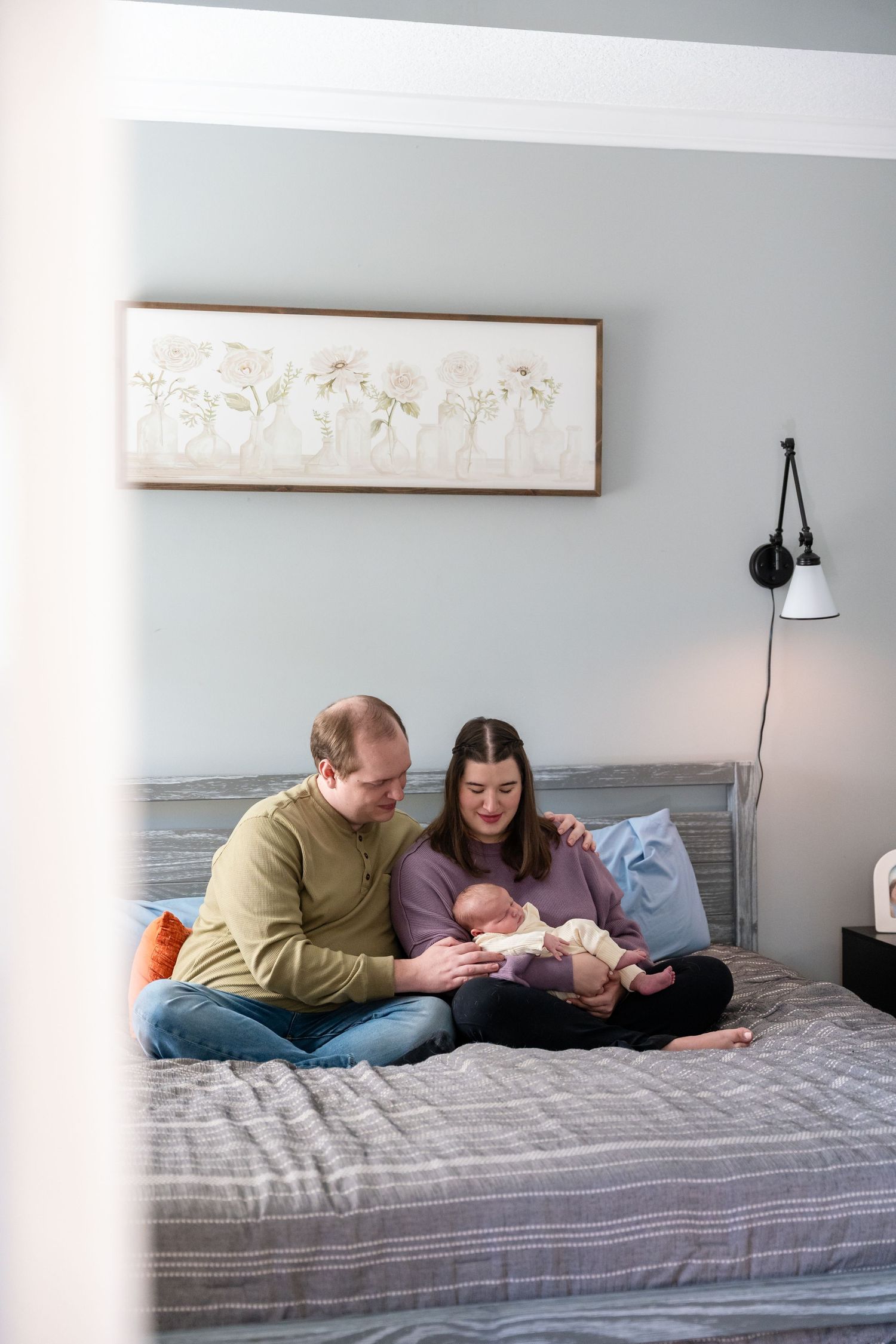 A couple sits on a bed holding their newborn baby in a softly decorated bedroom with blue-gray walls.