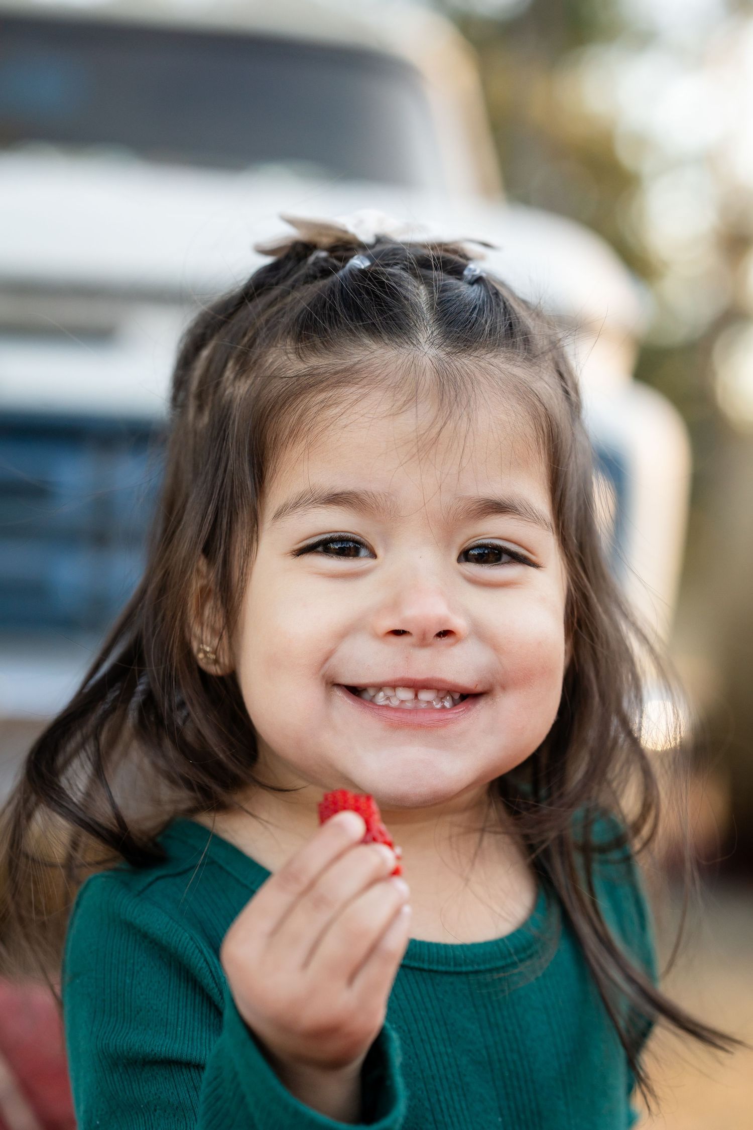 Small child in green sweater holding a strawberry while smiling outdoors.