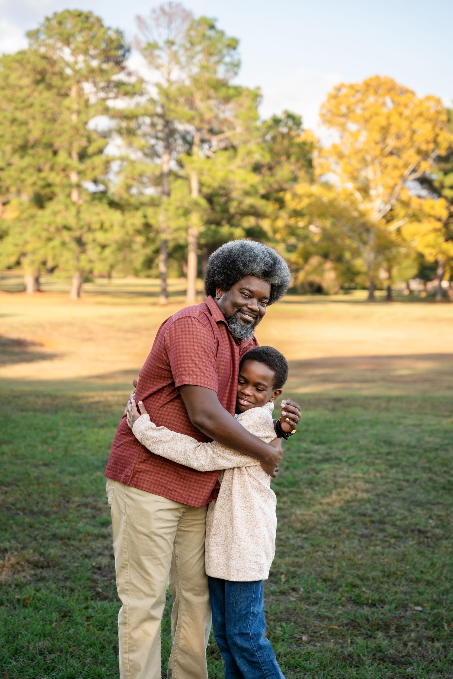 Two people sharing a warm embrace in a sunny park with trees in the background.