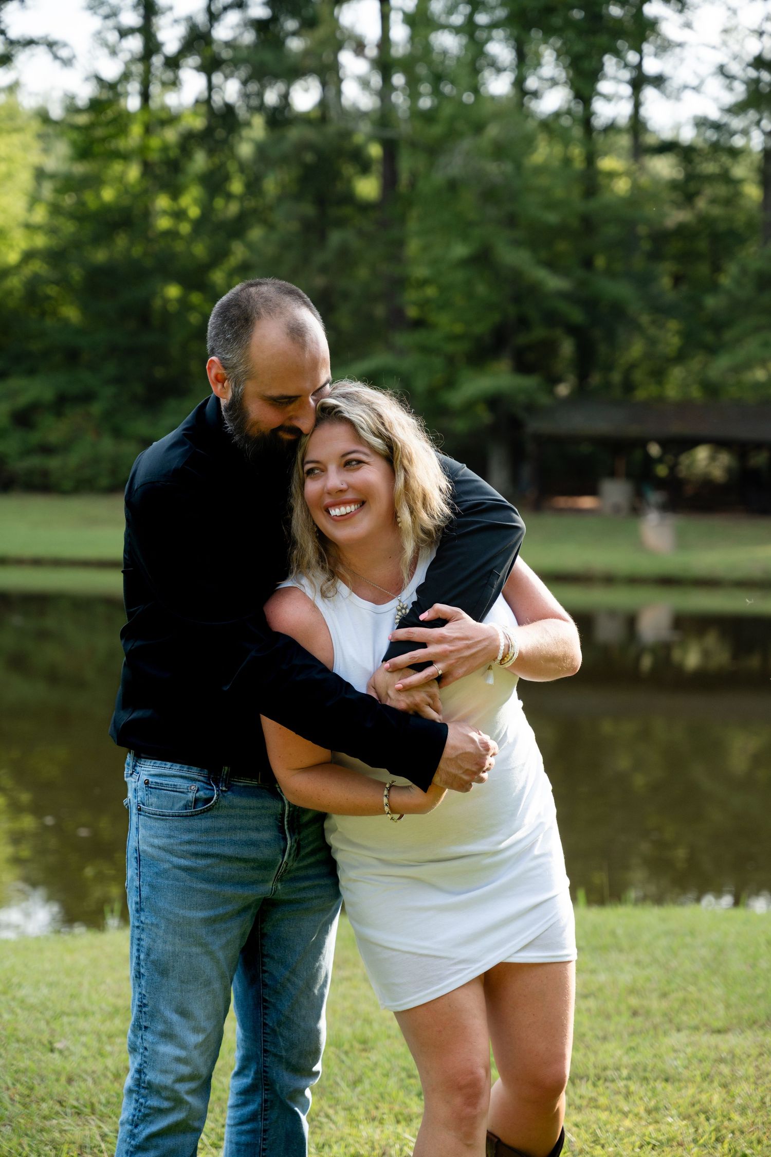 Happy couple laughing together by lake with wooden bridge in background.