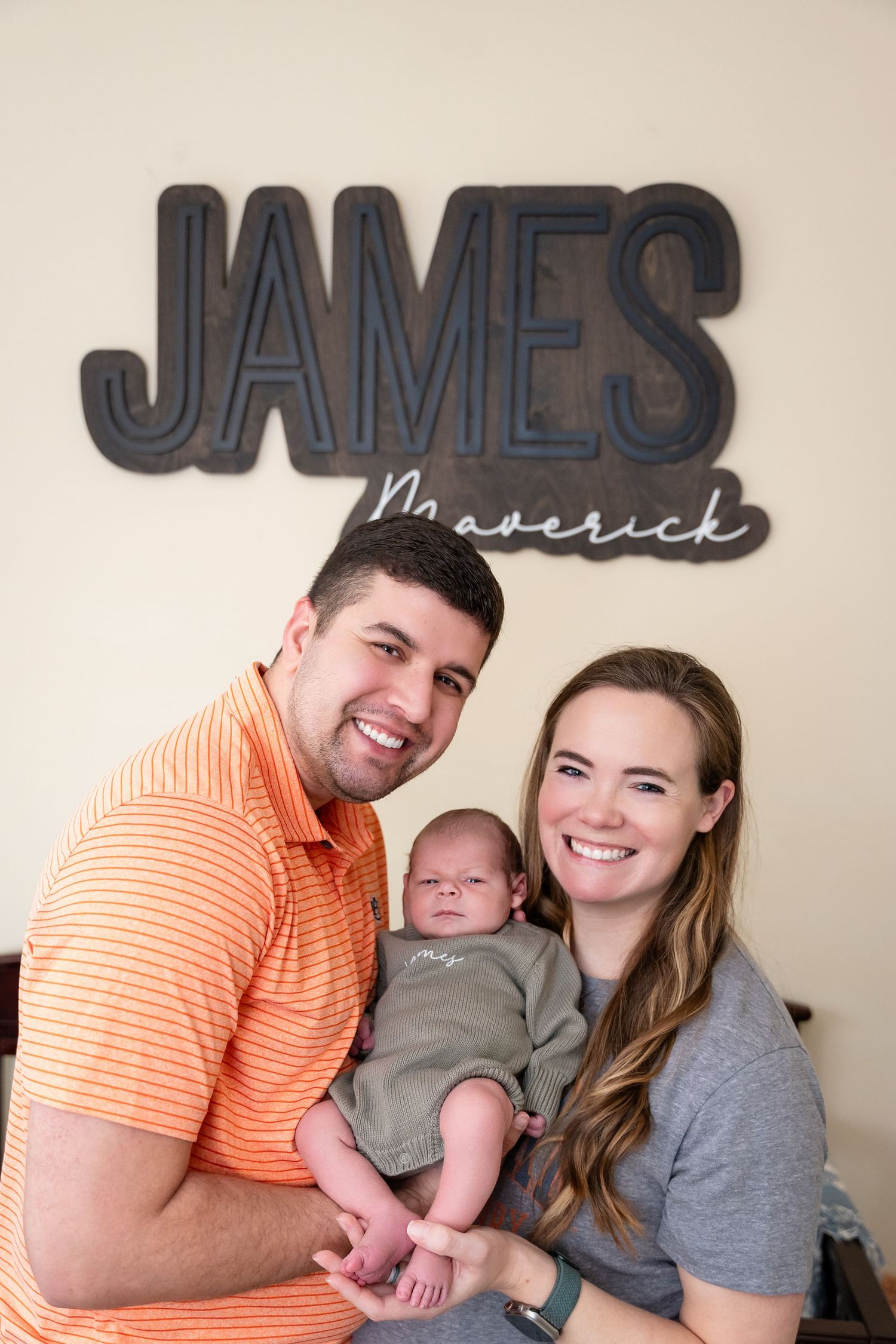 Family portrait under wall letters spelling 'JAMES' with baby in grey outfit.