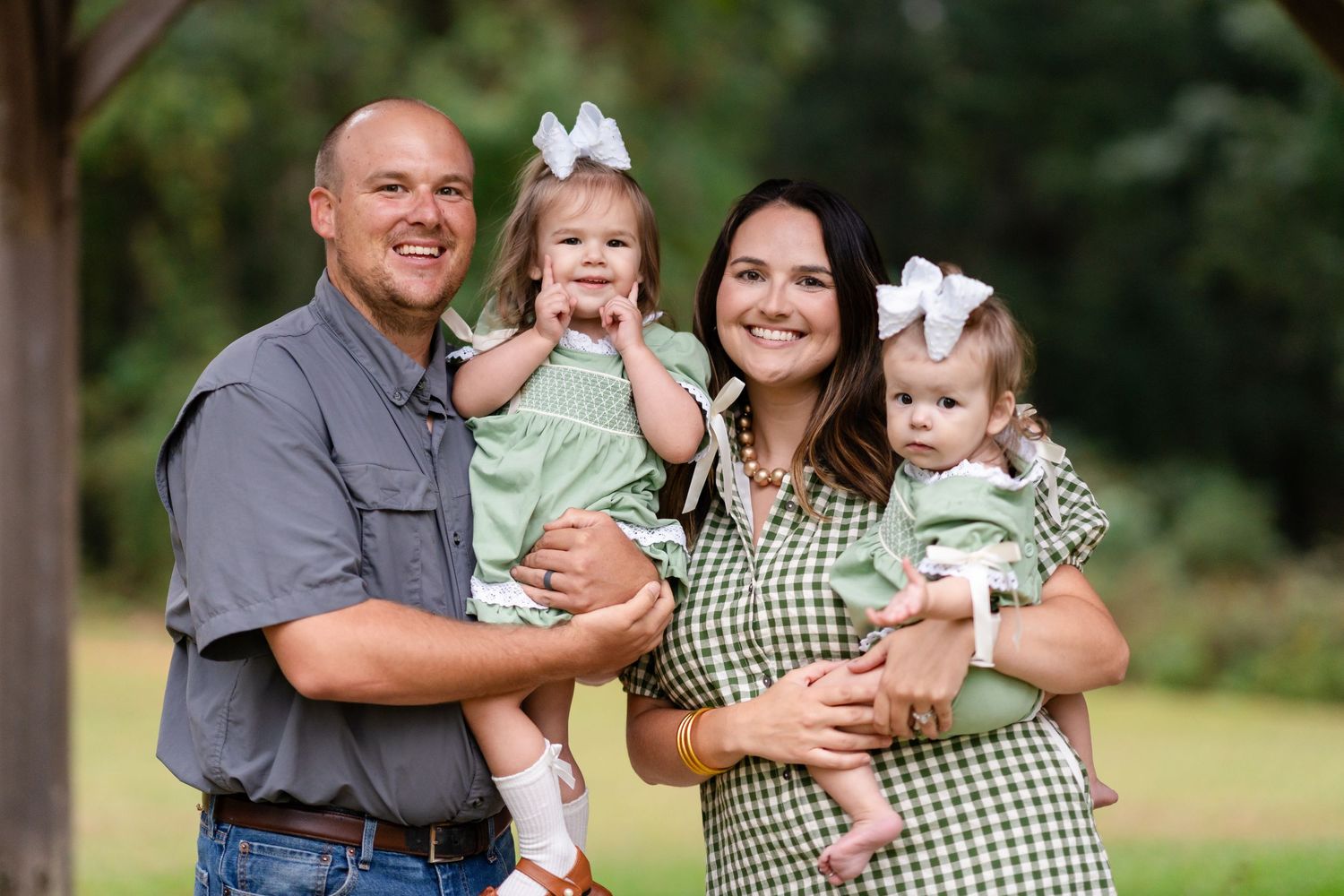 A smiling couple holds two toddler girls in matching green outfits and white hair bows during an outdoor family portrait in a grassy park.